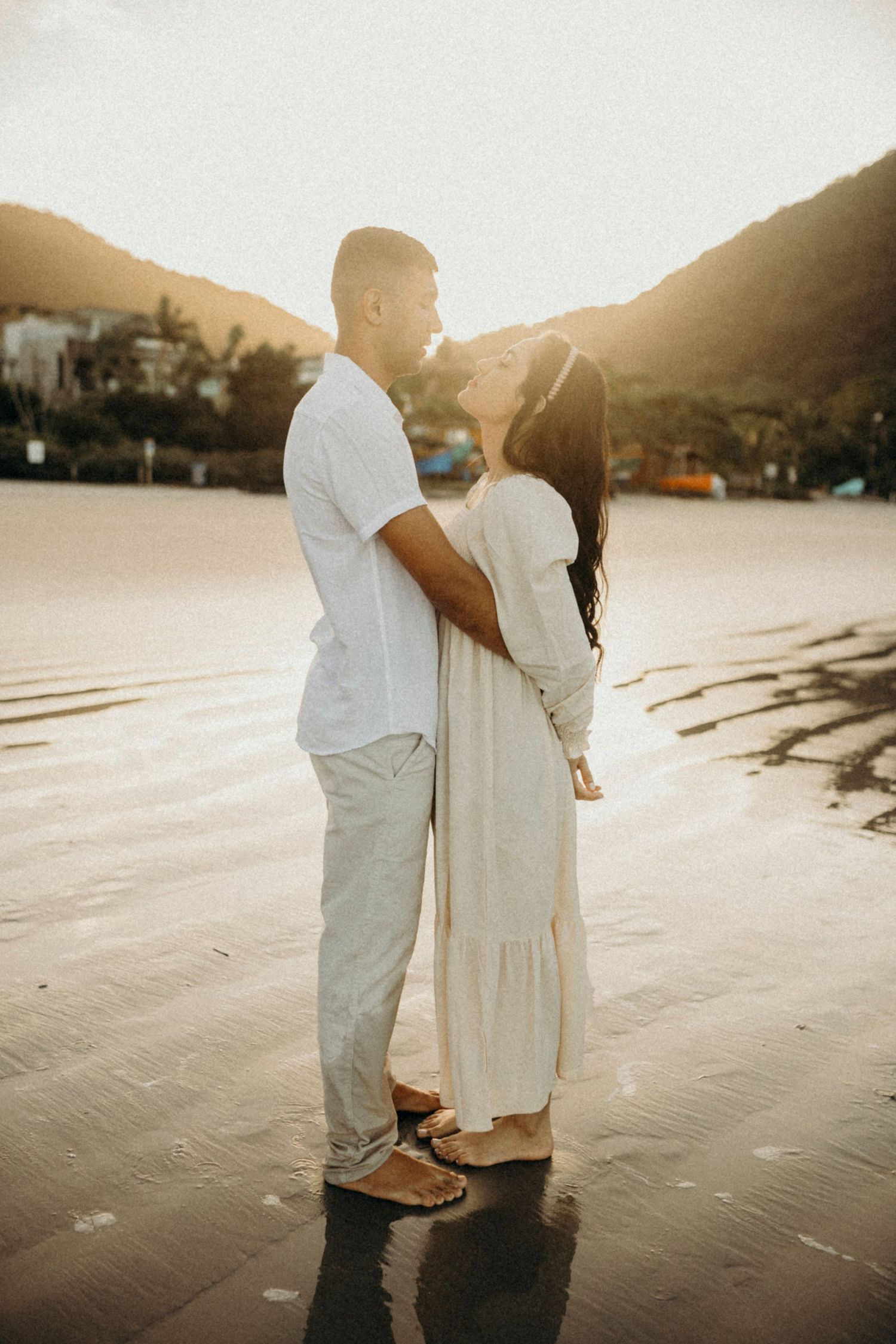 Couple in light colored clothing embrace on beach during golden hour.