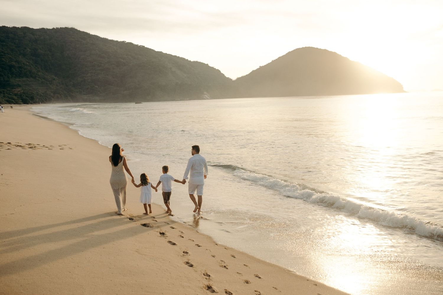 Family walking together along beach shoreline at golden sunset leaving footprints in sand.