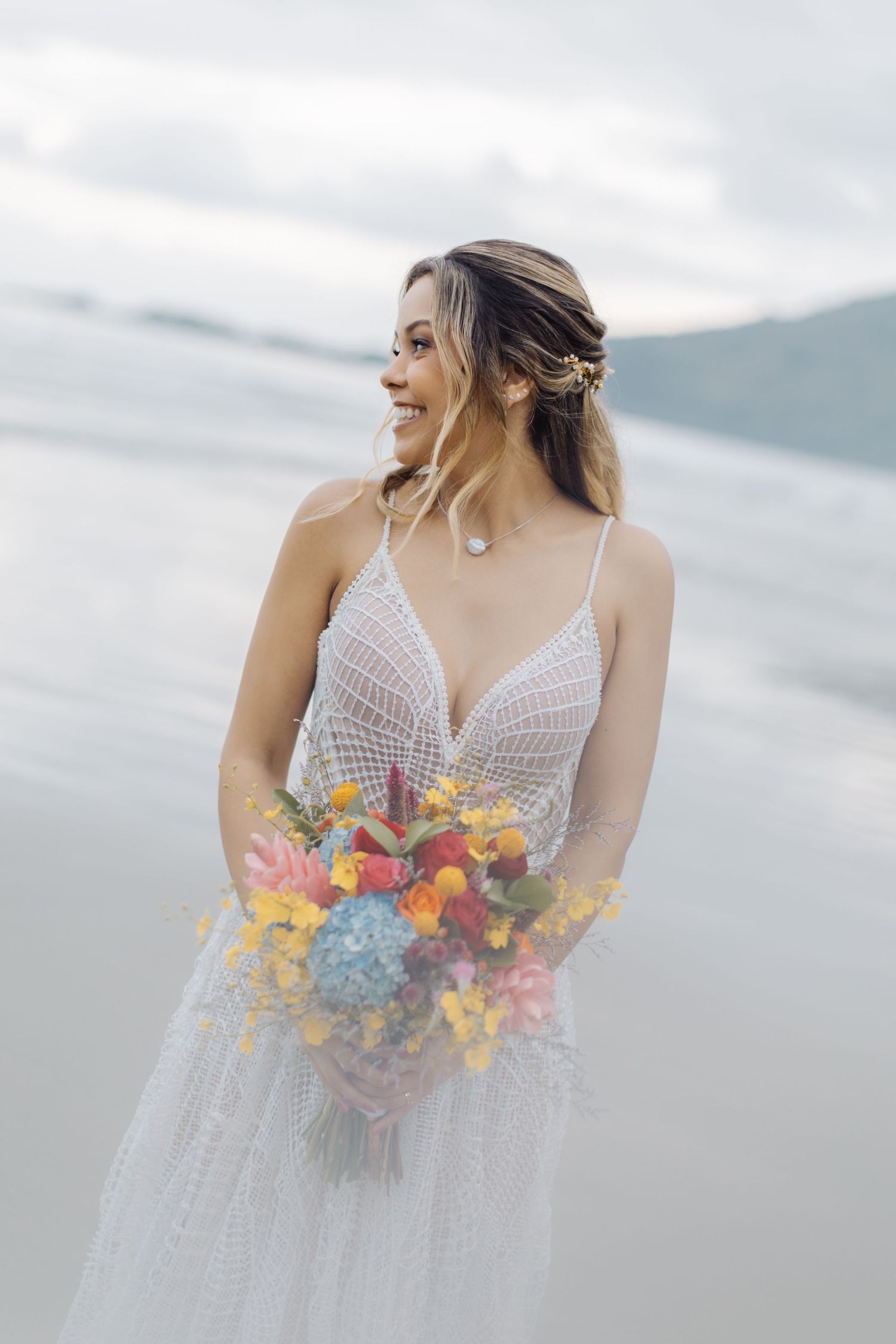 Bride in lace dress holding vibrant tropical bouquet against ocean backdrop.