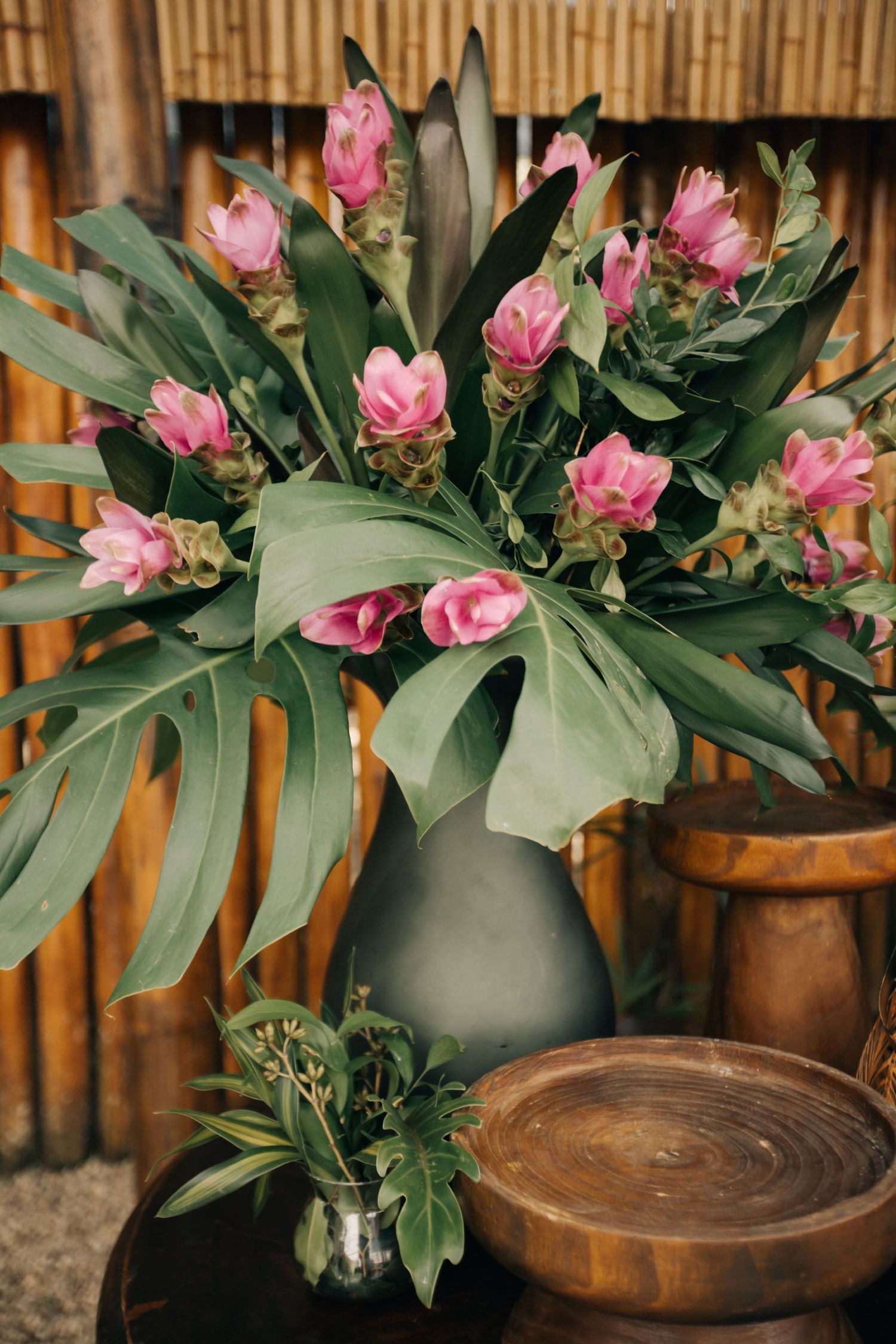 Pink ginger flowers arranged in a dark gray vase with tropical foliage against bamboo background.