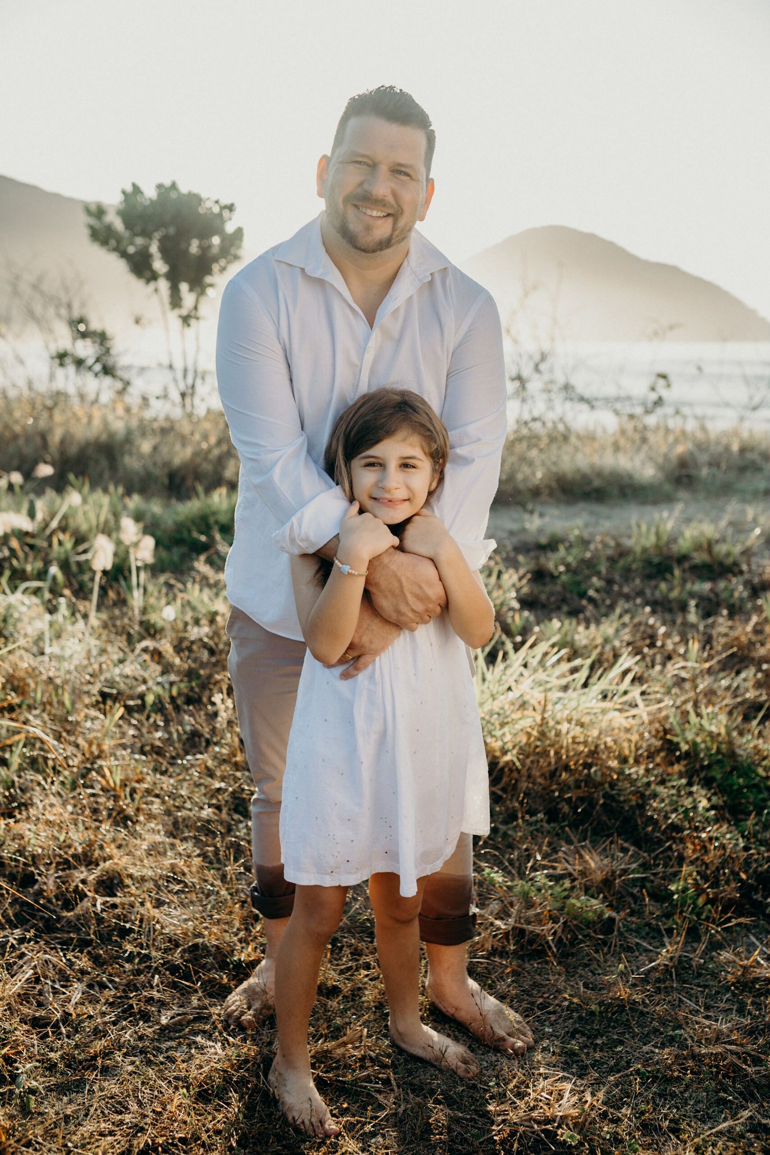 A touching outdoor portrait of a parent and child embracing in white attire against a scenic mountain backdrop.