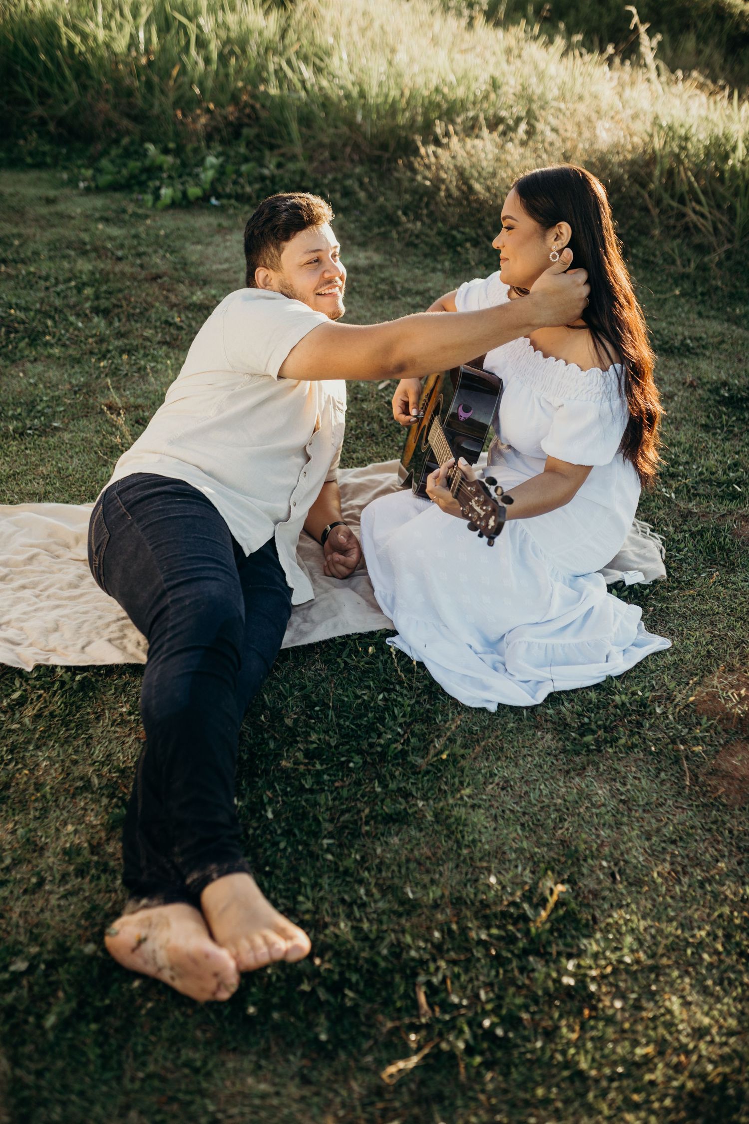 Playful couple enjoying outdoor music session with acoustic guitar on blanket.