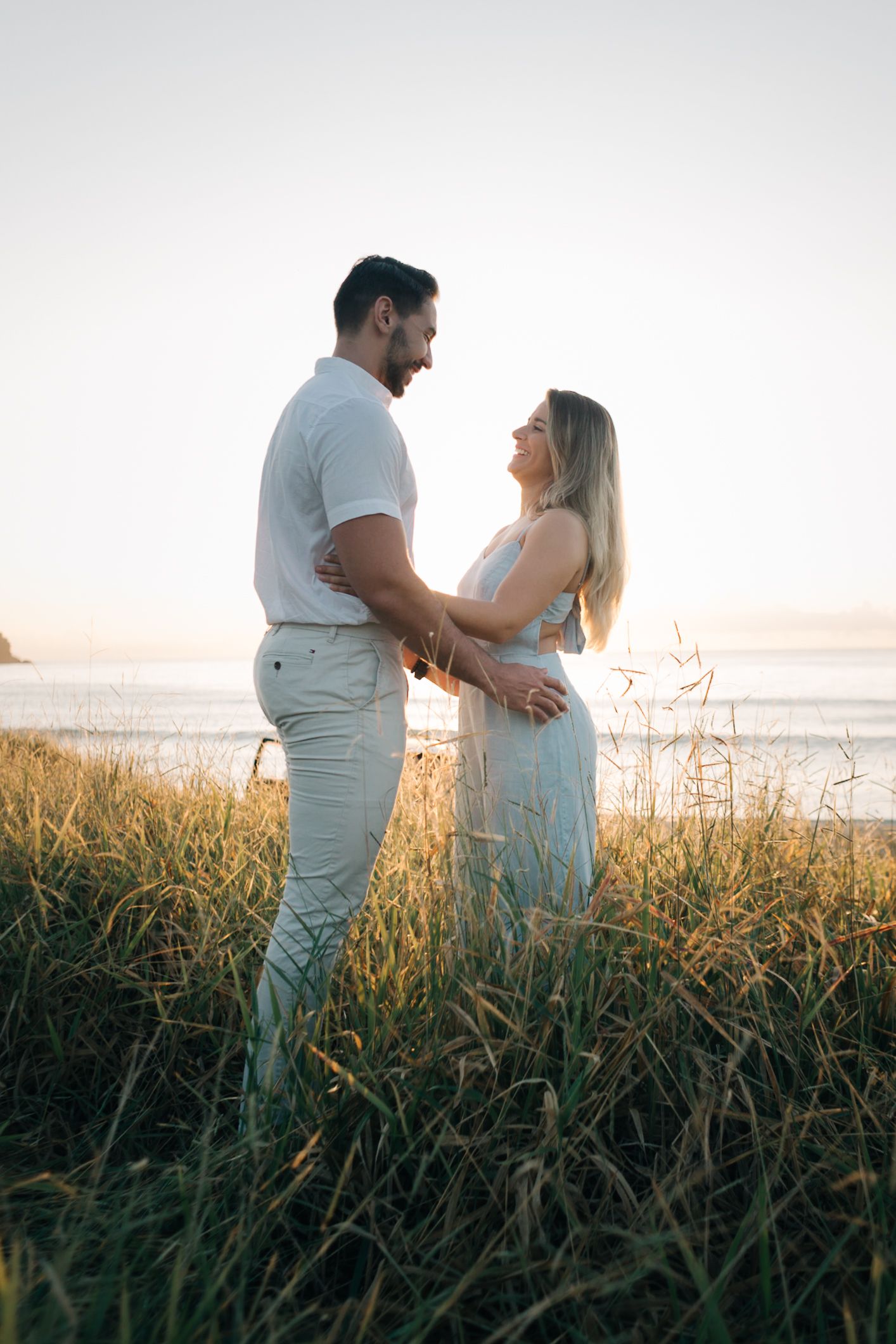 Couple dressed in white embracing in a seaside meadow during golden hour.