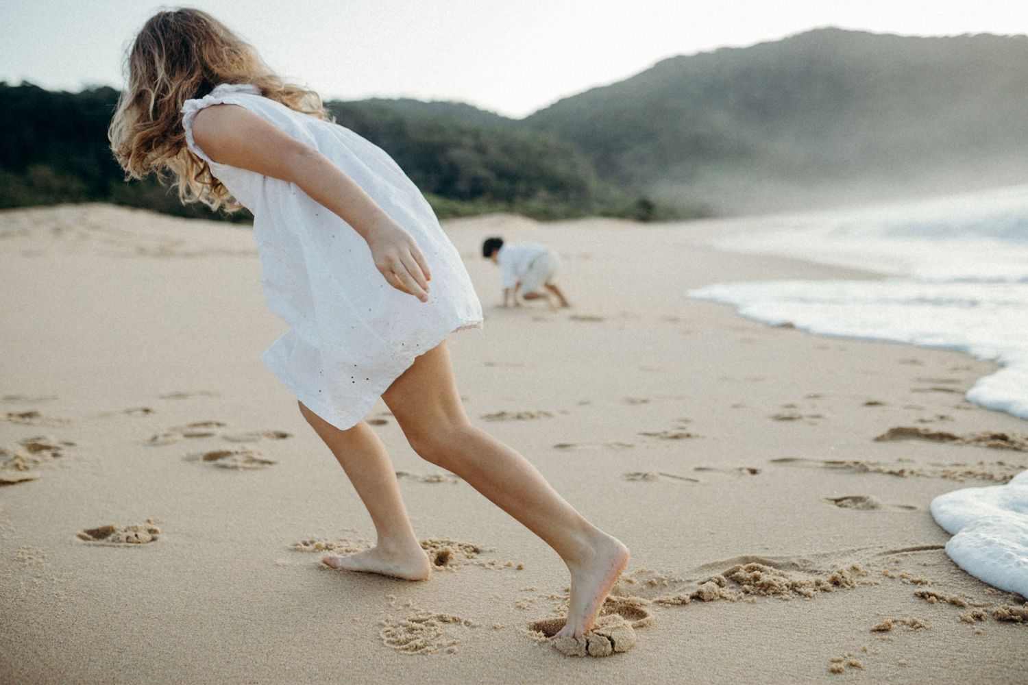 A person in a white dress running playfully on a sandy beach at sunset.