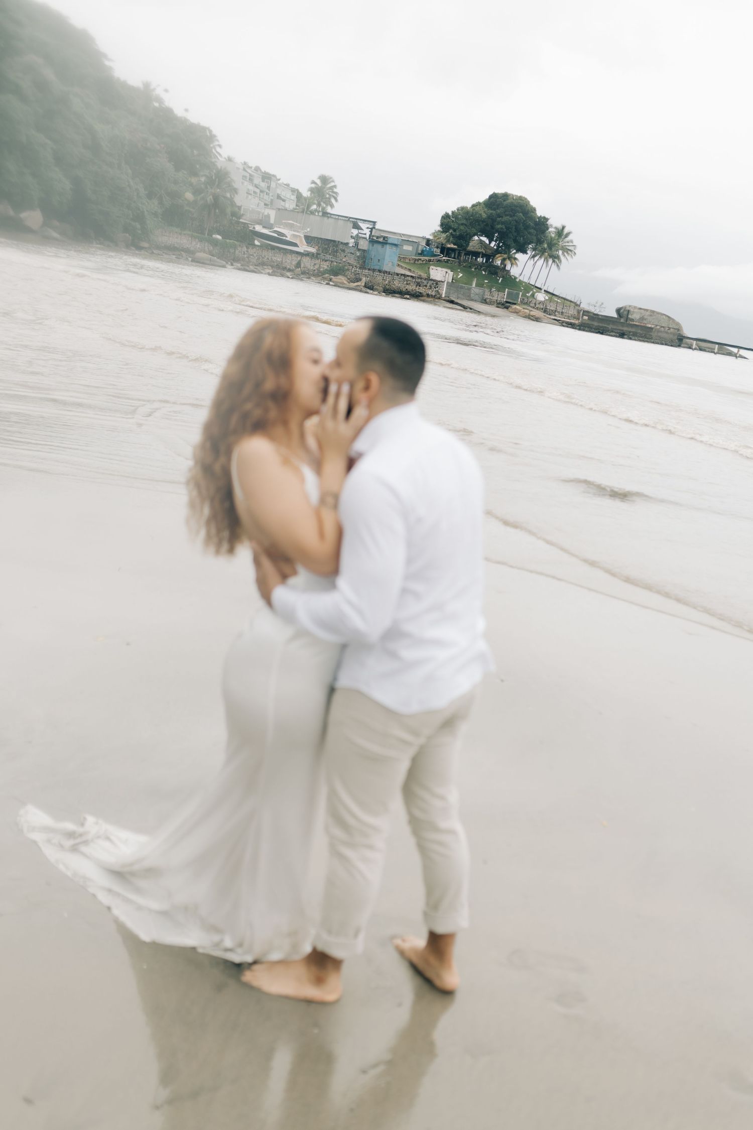 A dreamy beach photo captures an intimate moment between two people in white against a foggy backdrop.