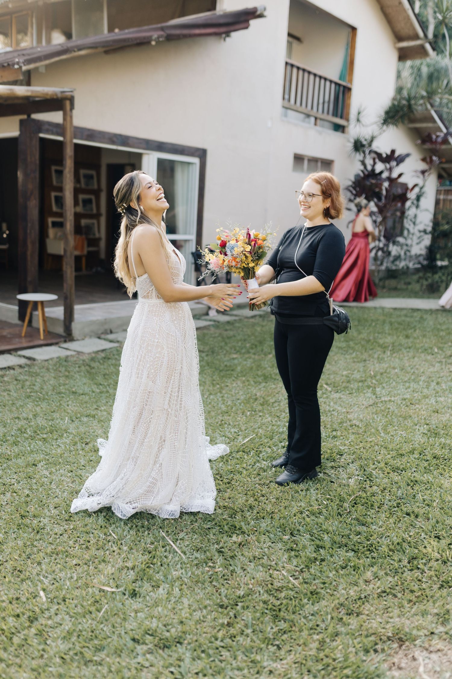 A bride in a flowy lace dress shares a joyful moment while receiving a colorful bouquet outside a house.