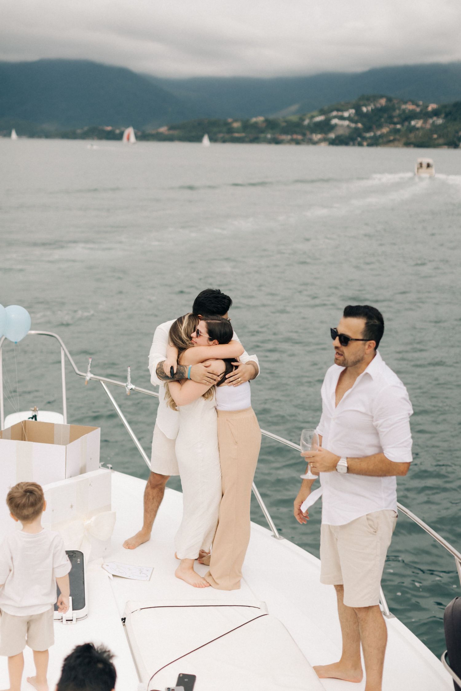 People embrace on a yacht deck with mountain views and sailboats in the distance.