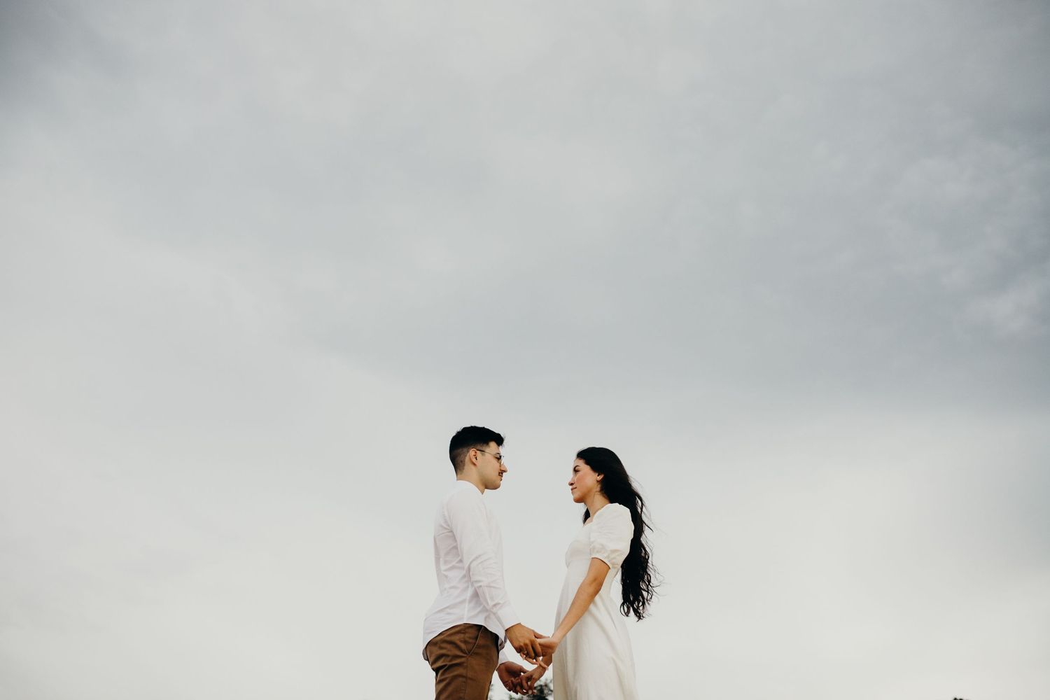 A romantic couple stands against an overcast sky in coordinated white and brown outfits.
