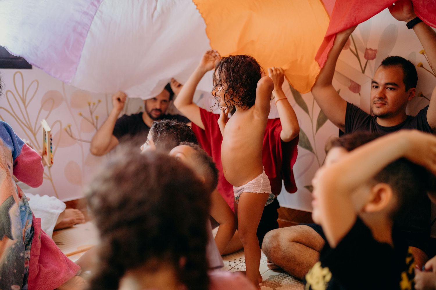 A group gathers under a vibrant parachute canopy during what appears to be a children's activity.
