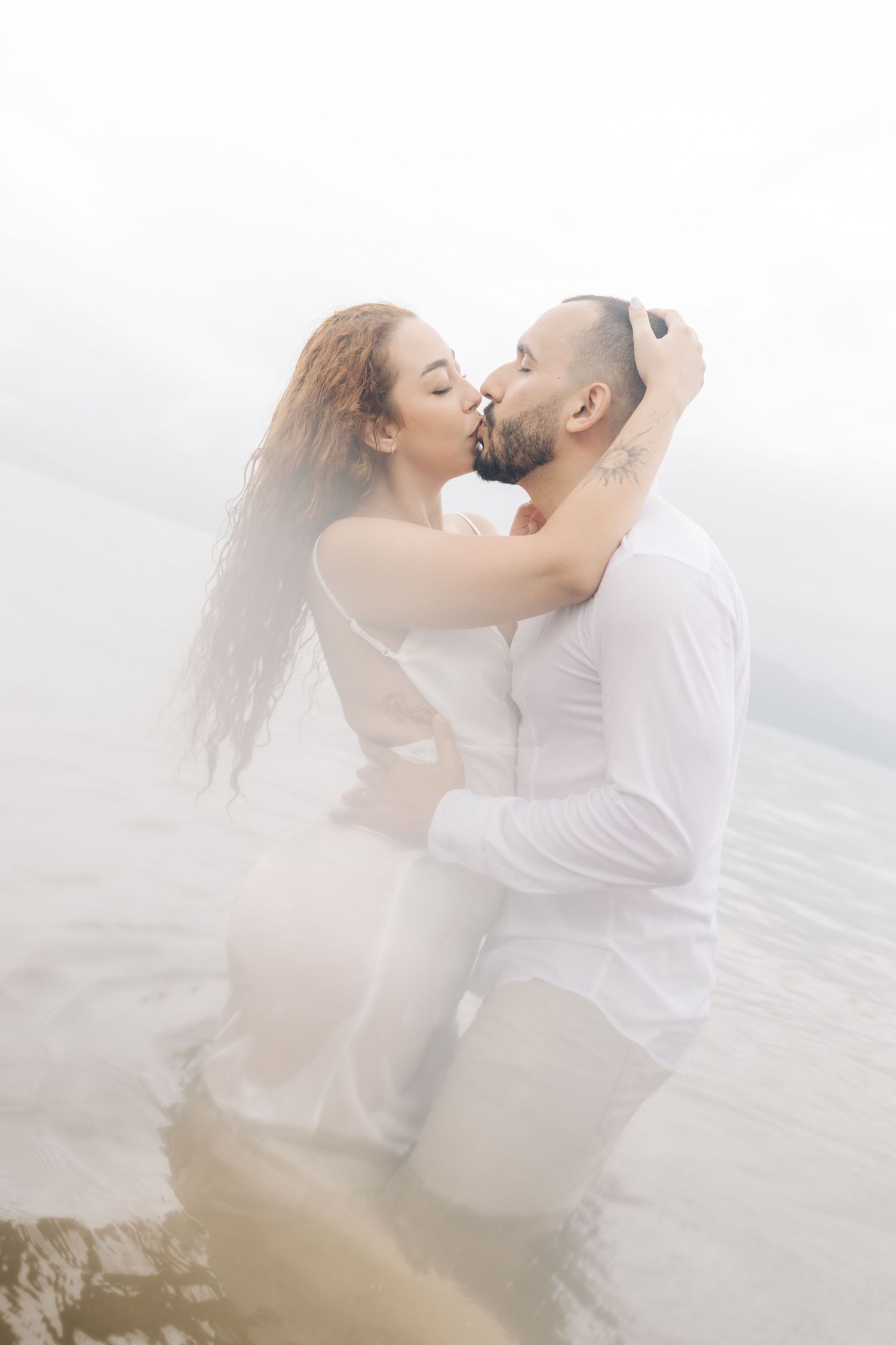 A dreamy beach photo of a couple in white embracing in misty ocean waters.