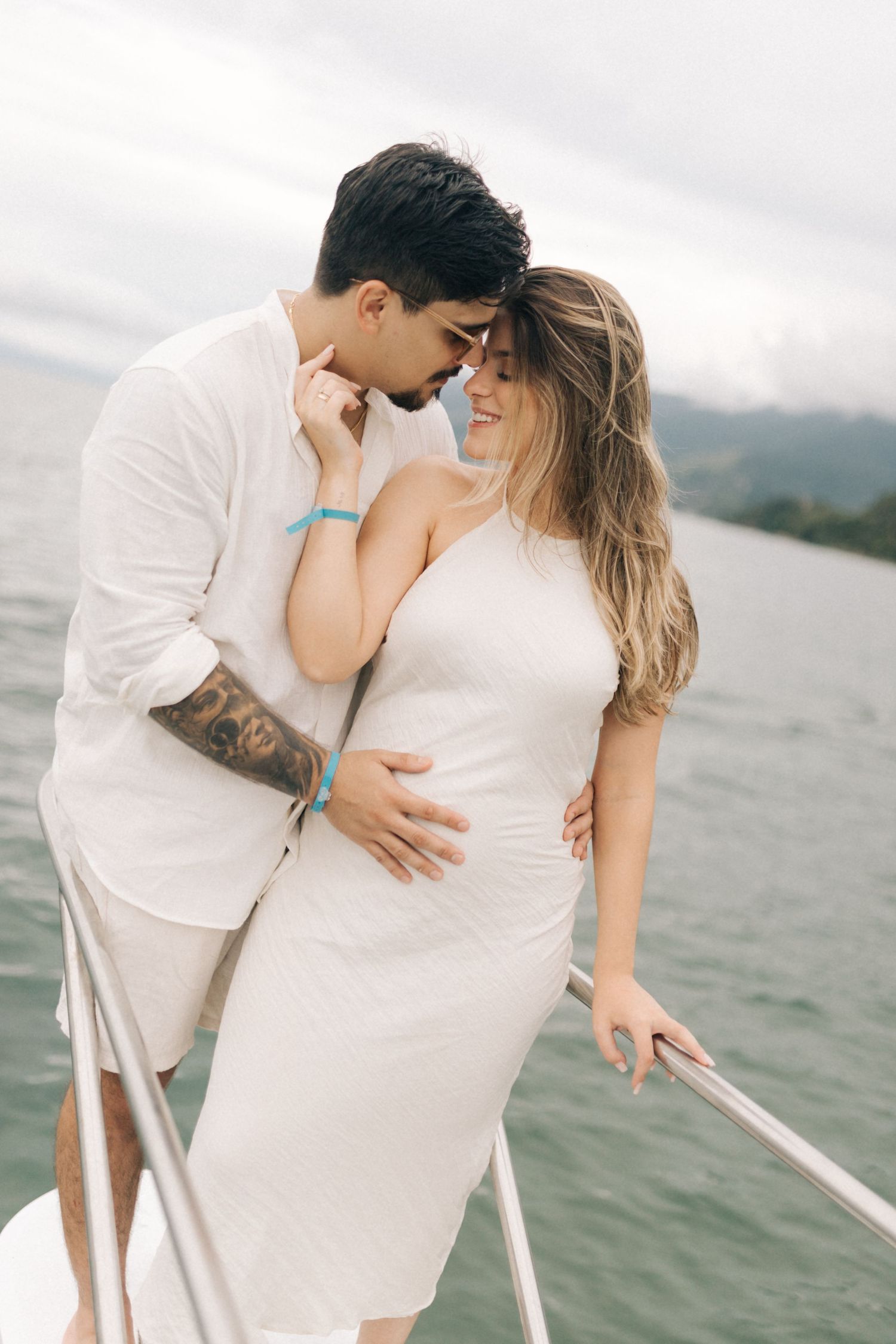 Loving couple in white outfits share intimate moment on boat with mountains in background.