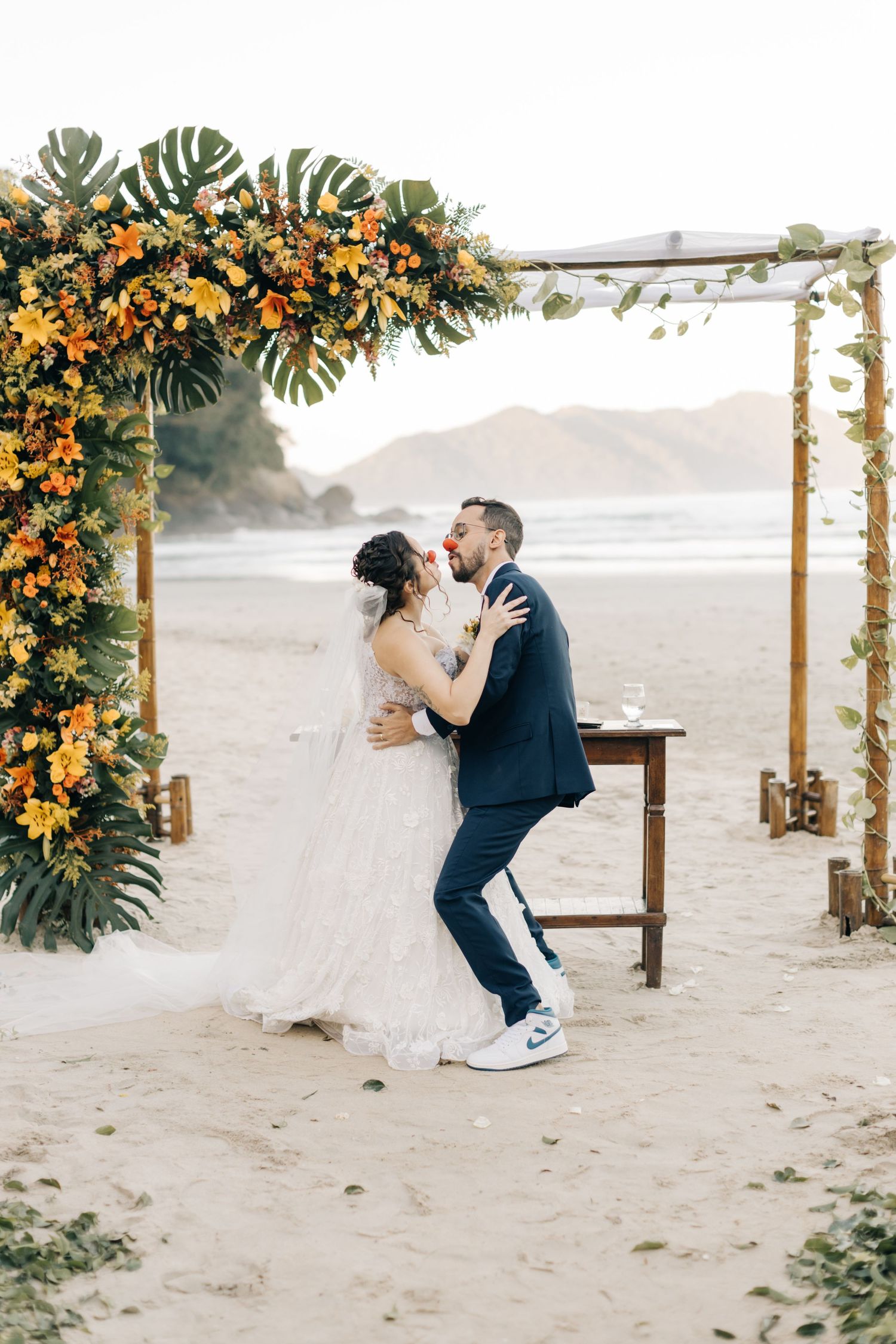Newlyweds embrace during their beach ceremony under an arch adorned with orange flowers and tropical leaves.