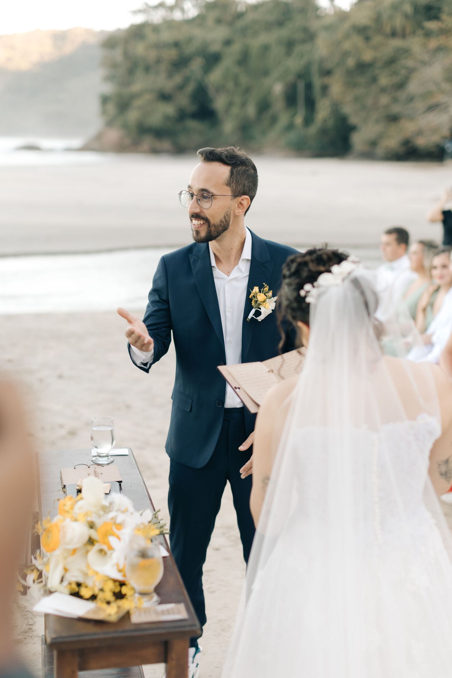 Groom in navy suit presides over beach wedding ceremony with yellow floral decorations.