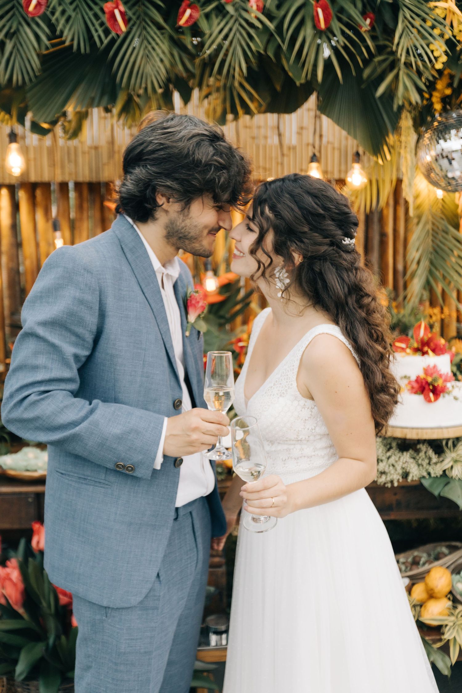 A romantic moment between newlyweds sharing champagne under tropical decor and string lights.