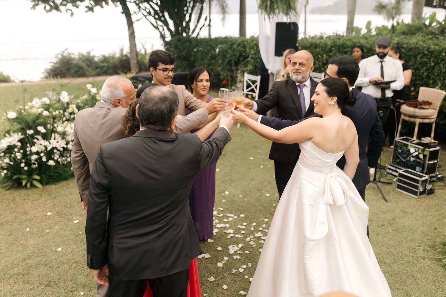 Wedding celebration toast with guests outdoors surrounded by flower petals on a grassy lawn.
