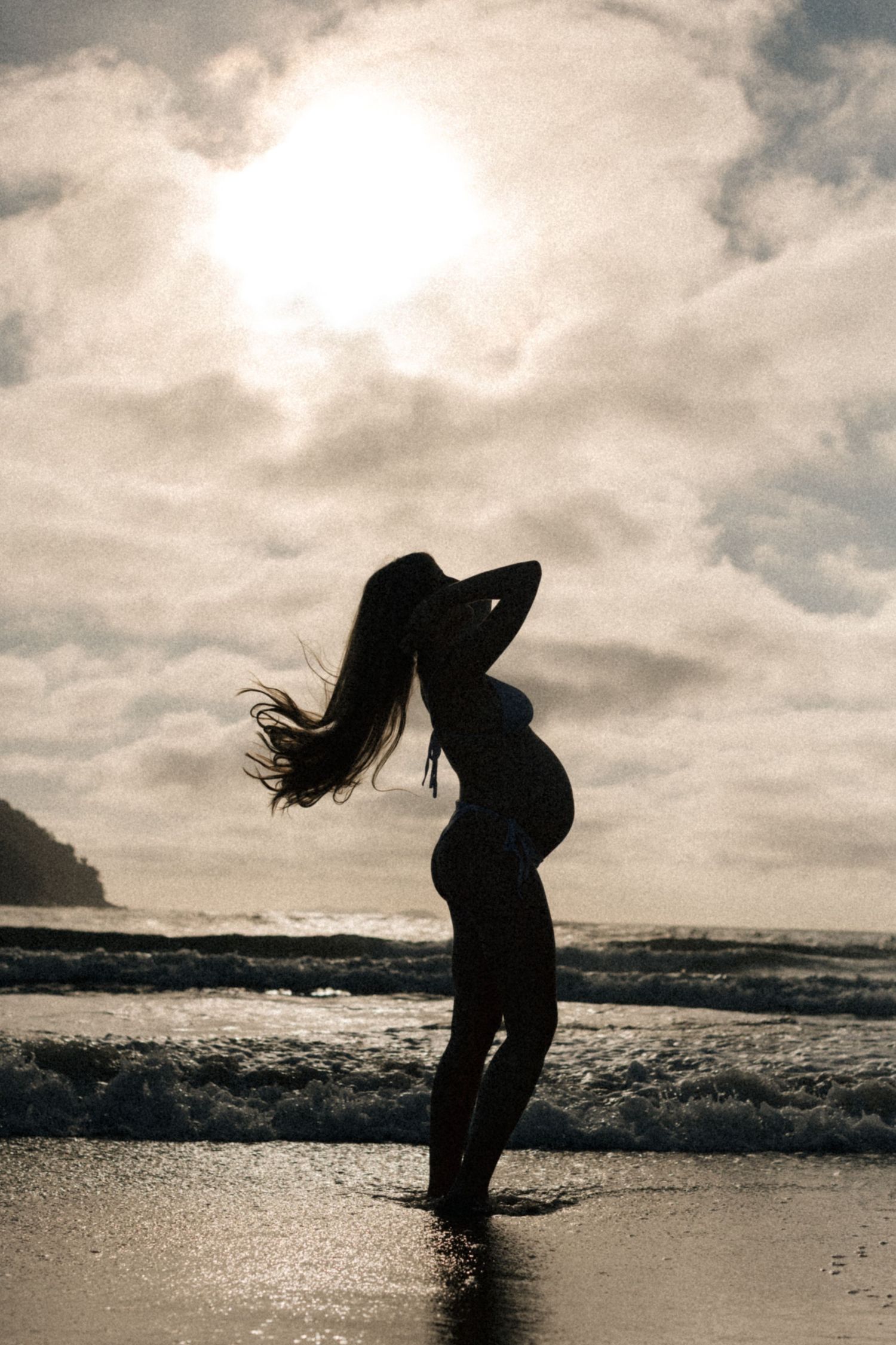 Silhouette of a pregnant woman arching backwards on a beach at sunset with dramatic cloudy sky.