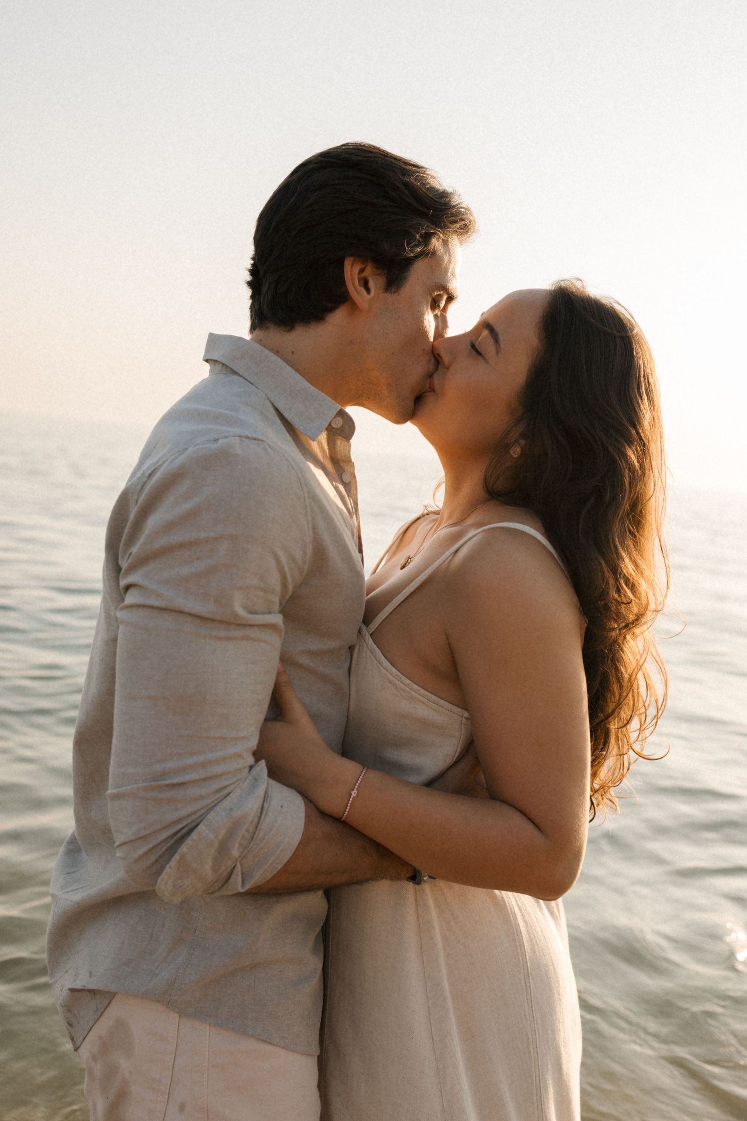 Romantic couple in light colored clothing share intimate moments by the ocean at sunset.