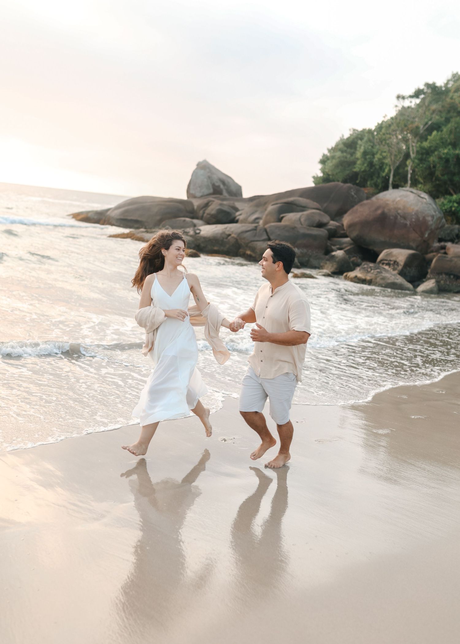 Two people in white clothing dance playfully along the shoreline with large rocks and ocean waves behind them.