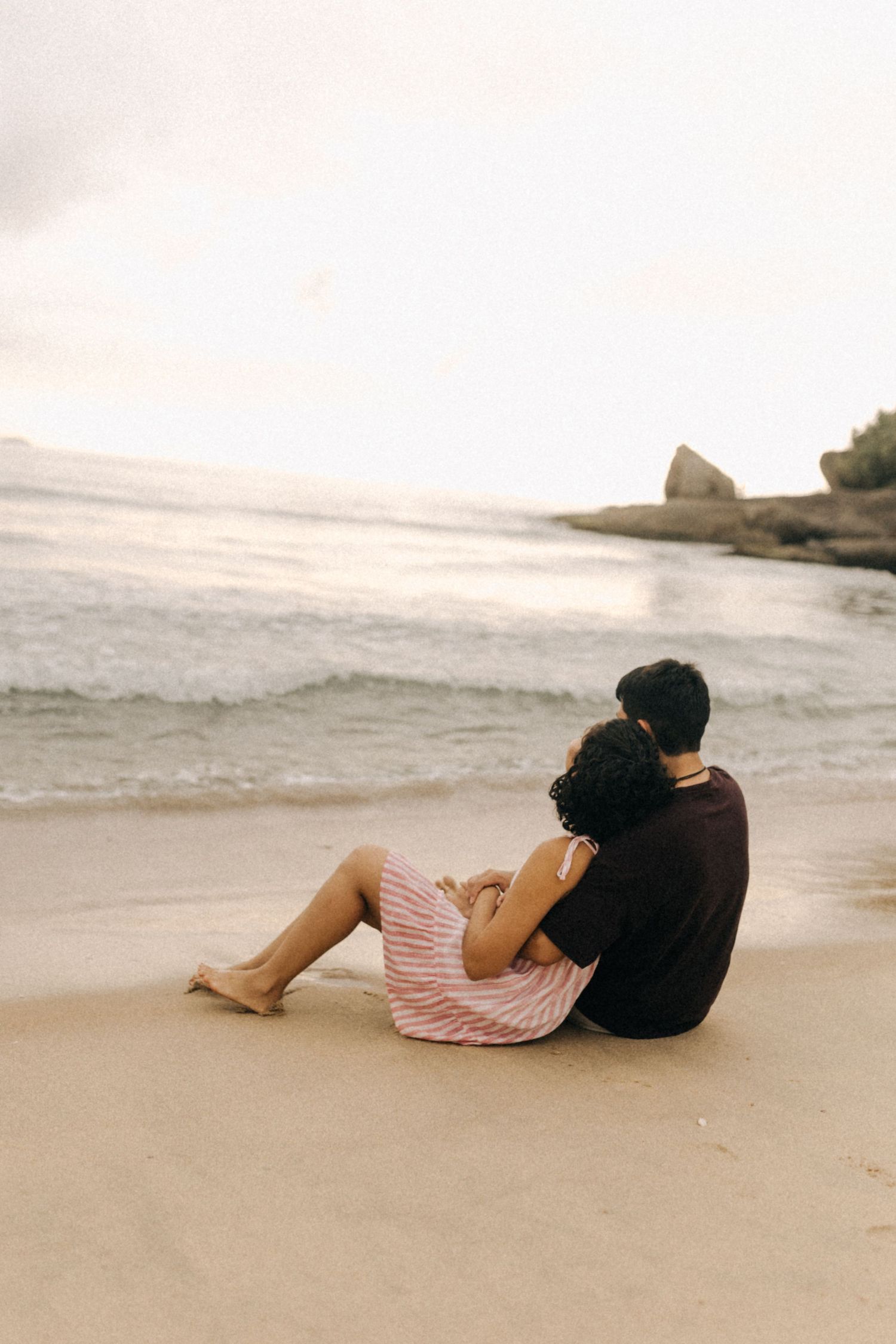 Um casal está sentado em uma praia tranquila ao pôr do sol, com ondas do mar e falésias rochosas ao fundo.