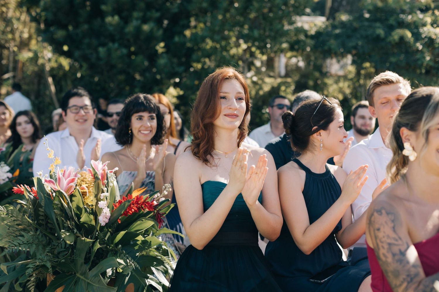 Wedding attendees in formal attire applaud during an outdoor celebration with floral decorations.