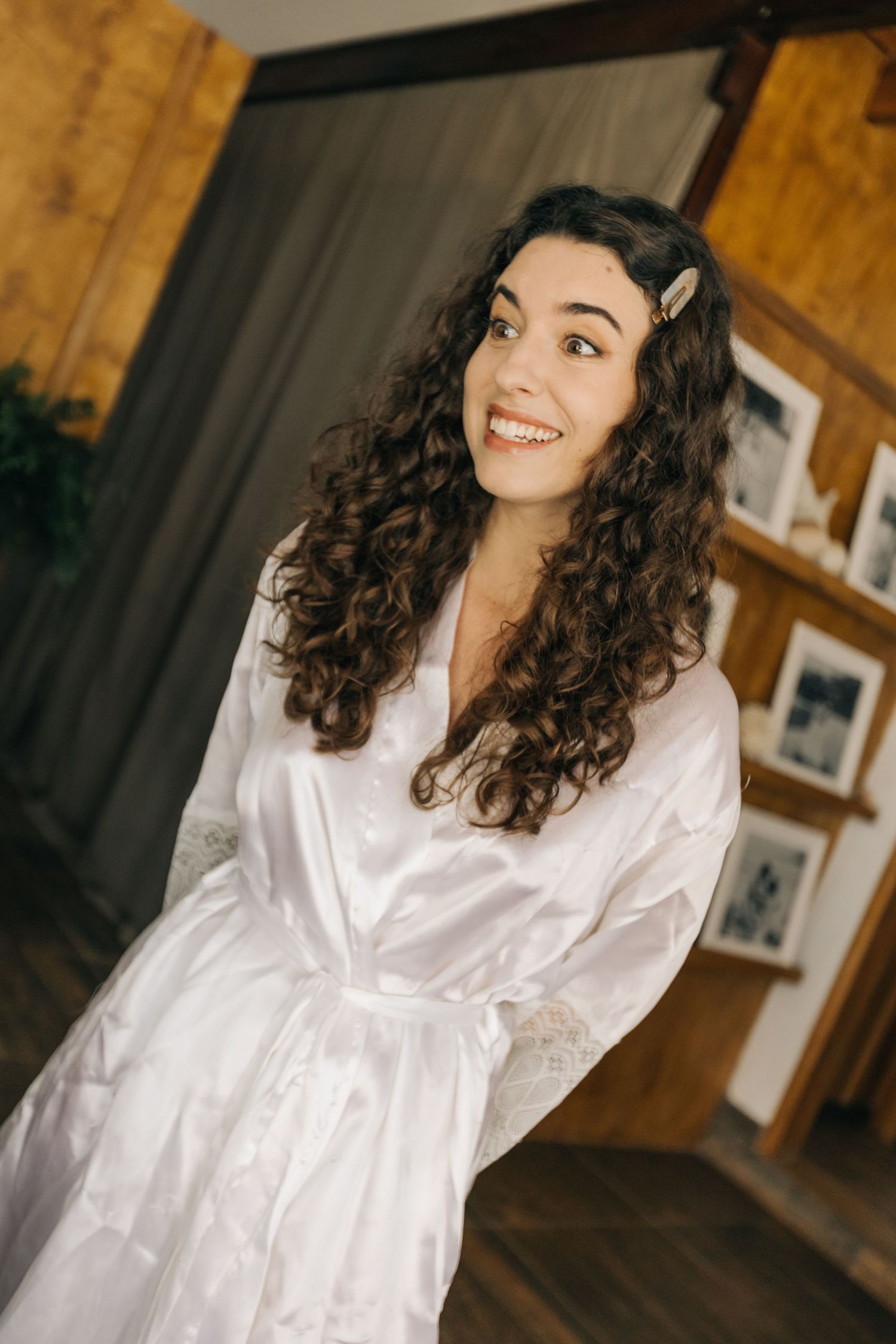 Woman in white silk robe with curly dark hair and hair clips smiling in a rustic indoor setting.