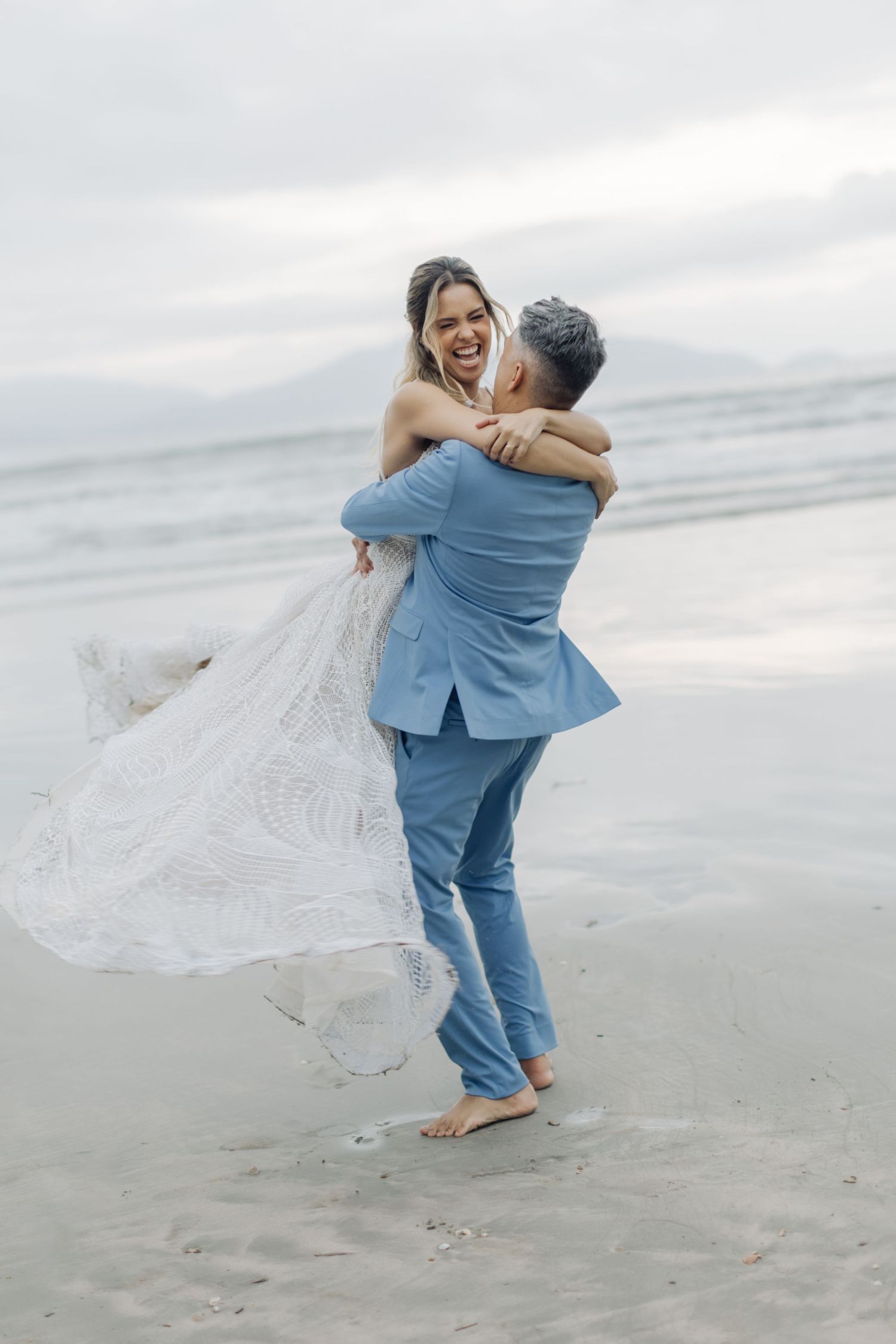 Romantic beach photoshoot with couple in blue suit and white lace dress dancing in the sand.