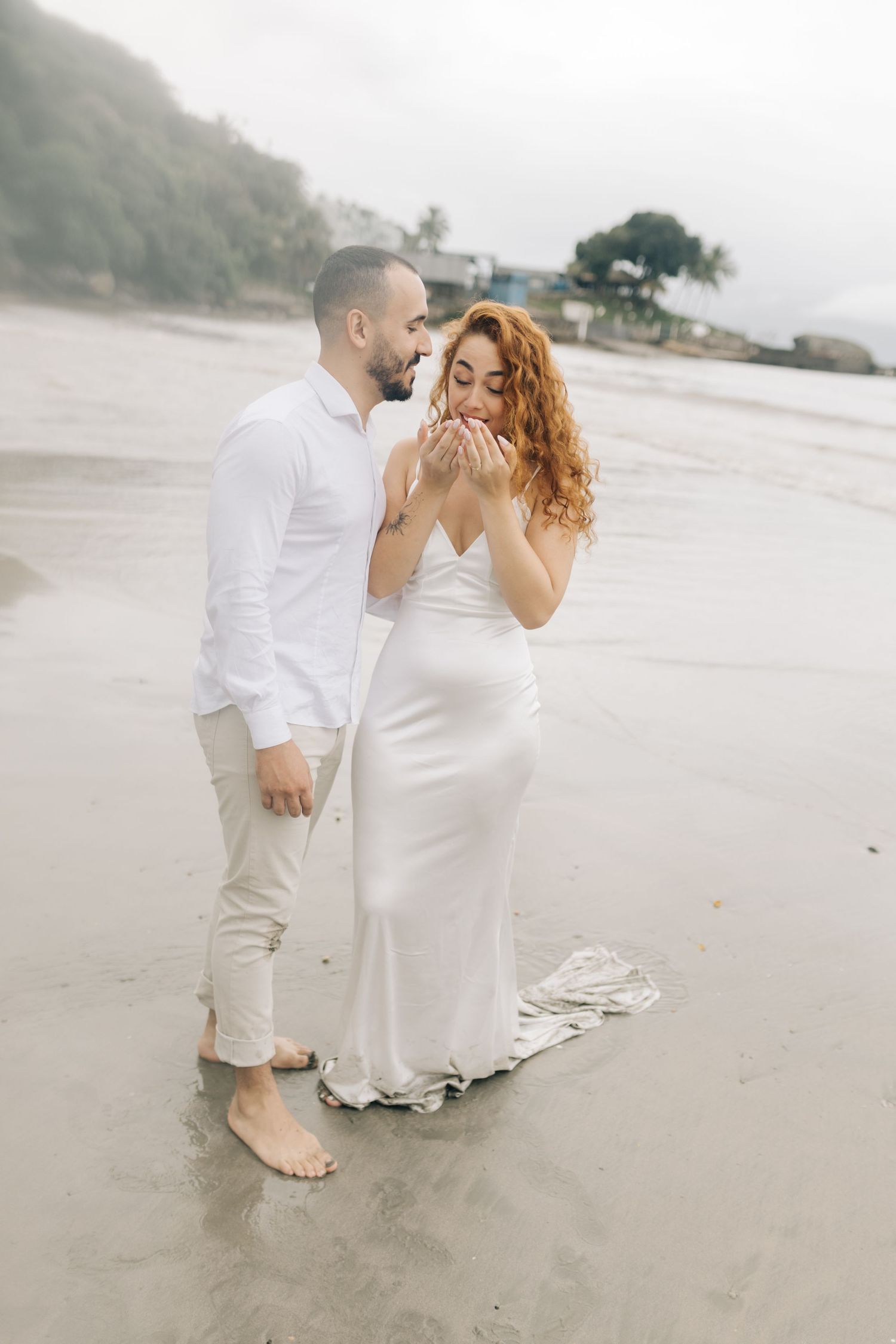 Romantic beach proposal scene with couple in white attire standing in shallow water.