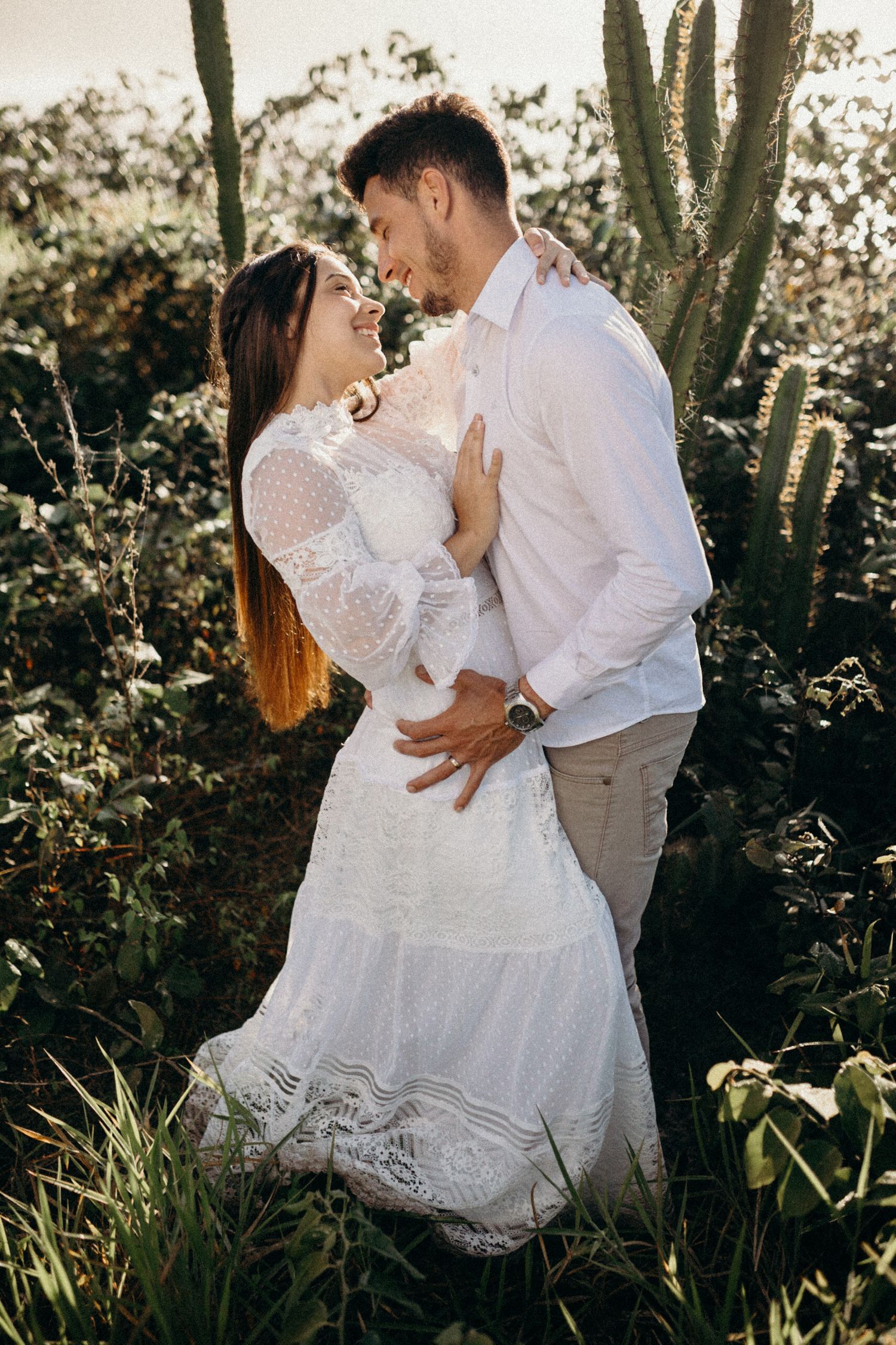 Couple in white and beige share tender moment among desert cacti and wild grass.