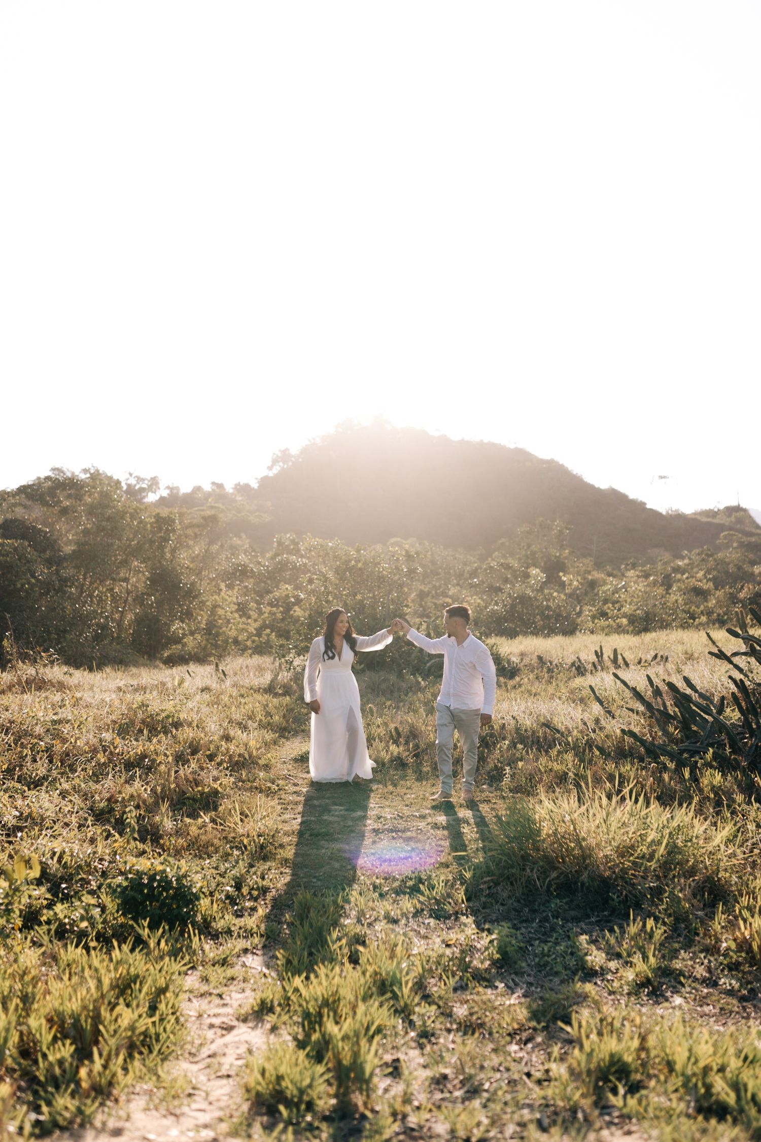 A couple in white outfits walks hand in hand through a sunlit mountain trail.