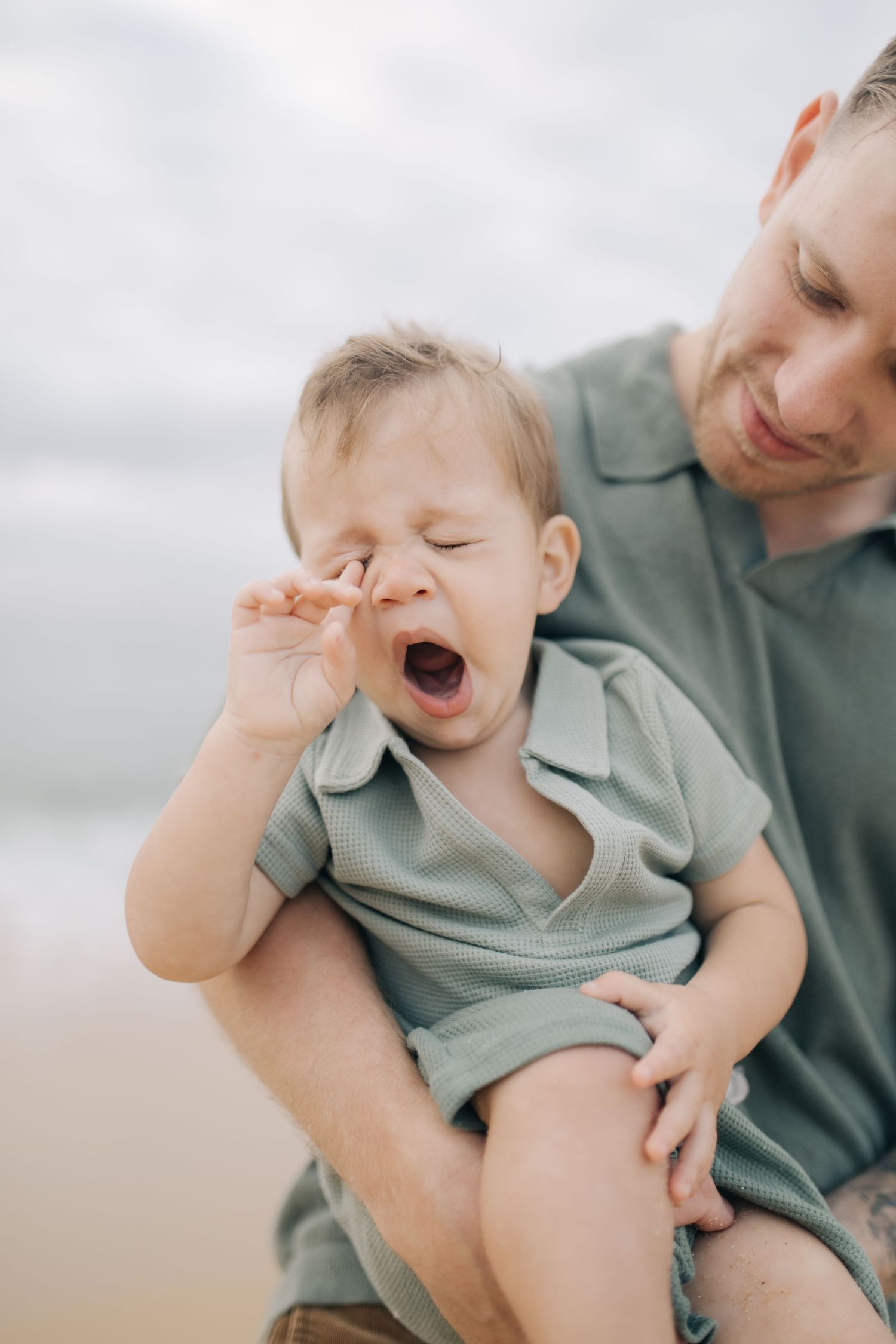 A toddler rubs their eyes while being held during a beach photoshoot.