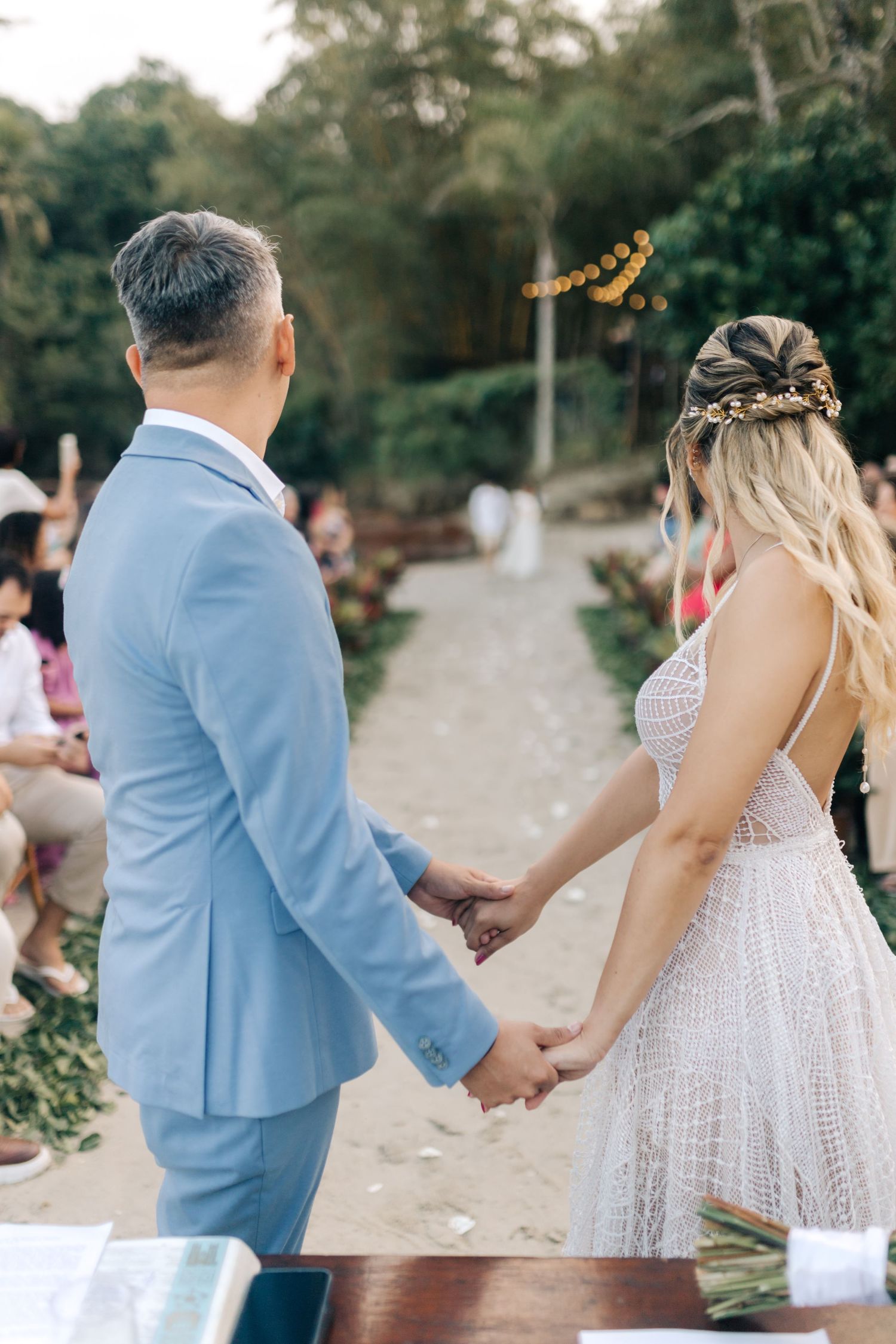 Couple holds hands during outdoor wedding ceremony with decorative lighting and foliage.