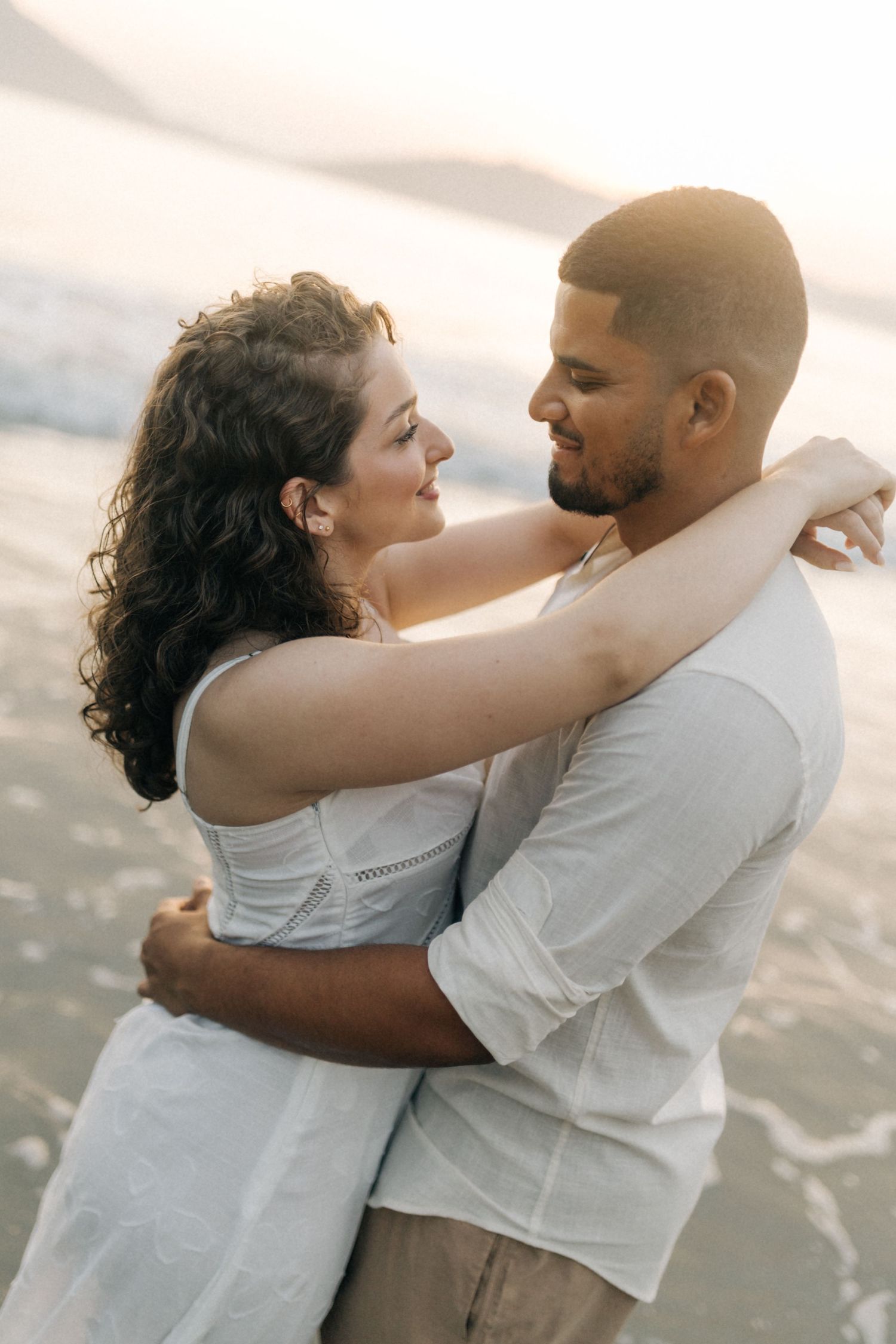 Intimate portrait of couple in white attire embracing at beach during sunset.