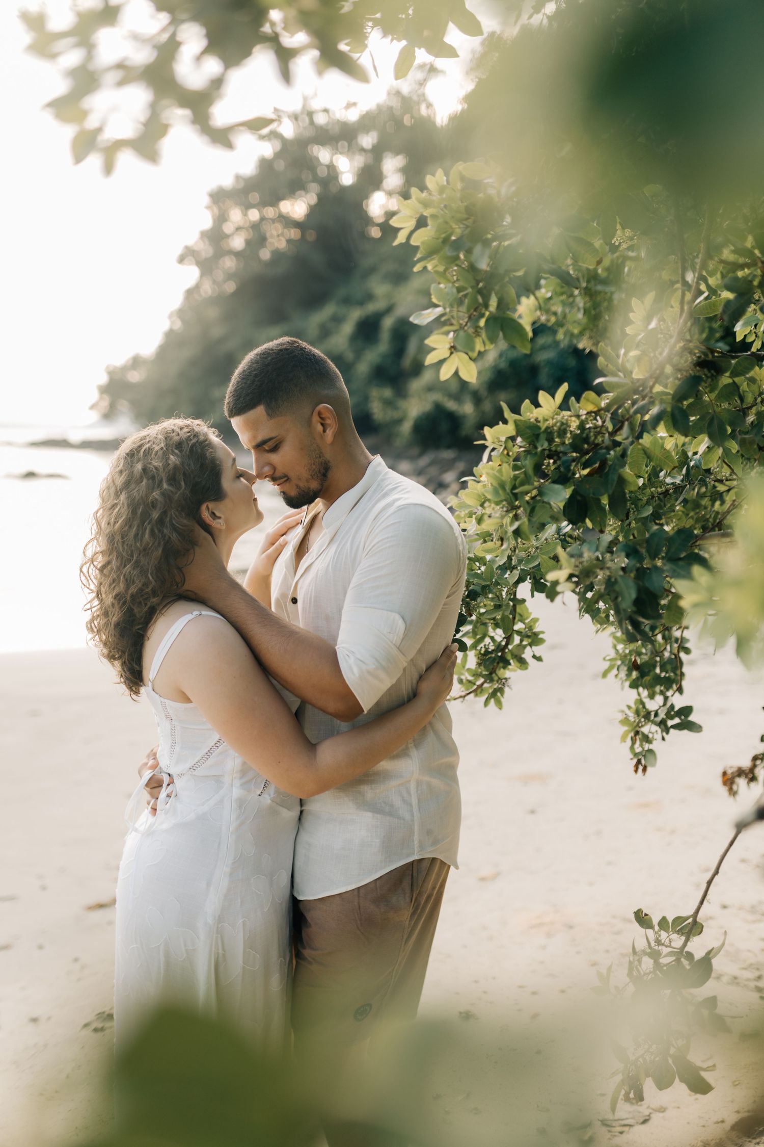 Romantic couple embracing on beach with leafy foliage in foreground.