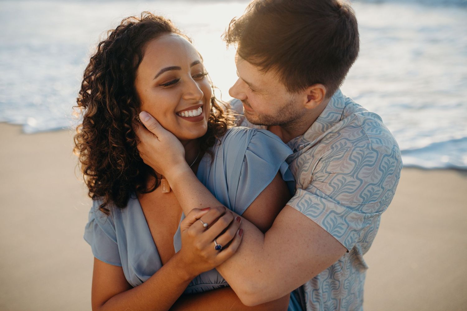 Joyful couple laughing and embracing on beach with ocean waves behind them.