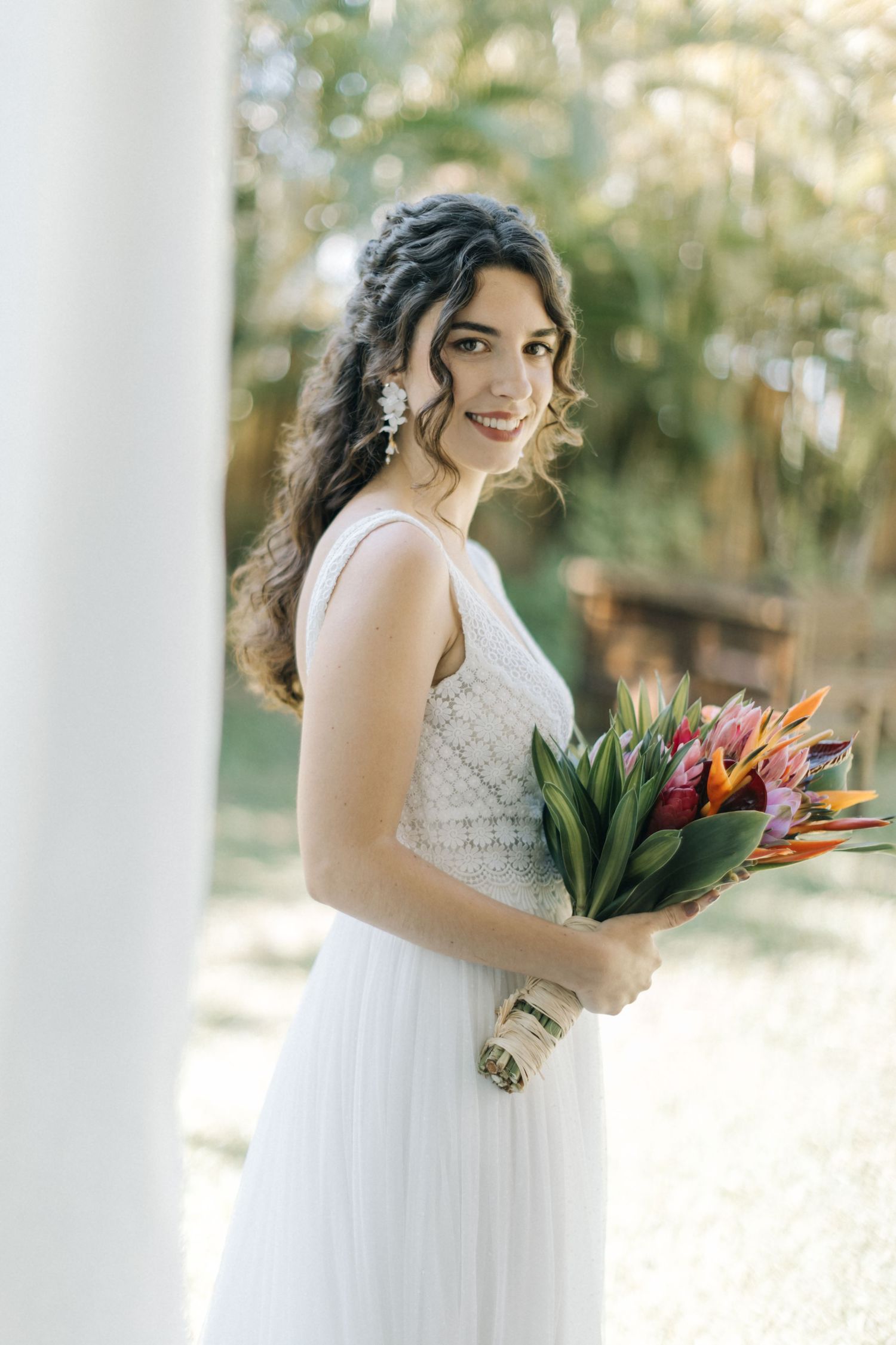 Elegant bride carrying tropical flowers poses on white pillared porch.