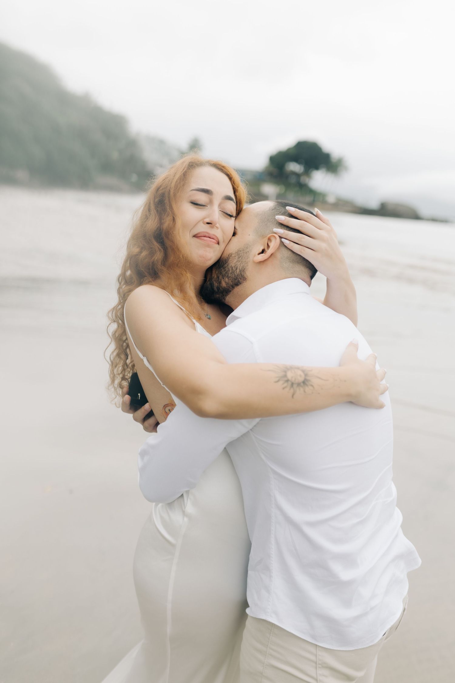 A tender moment between two people in white clothing on a cloudy beach with mountains in the background.