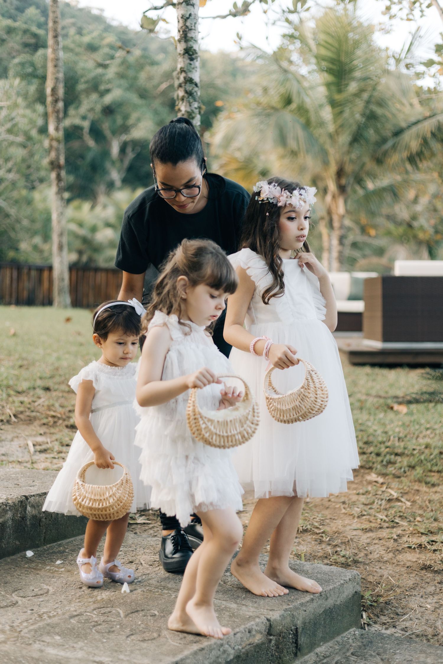 Three children in white dresses carry flower baskets while walking with an adult at an outdoor event.
