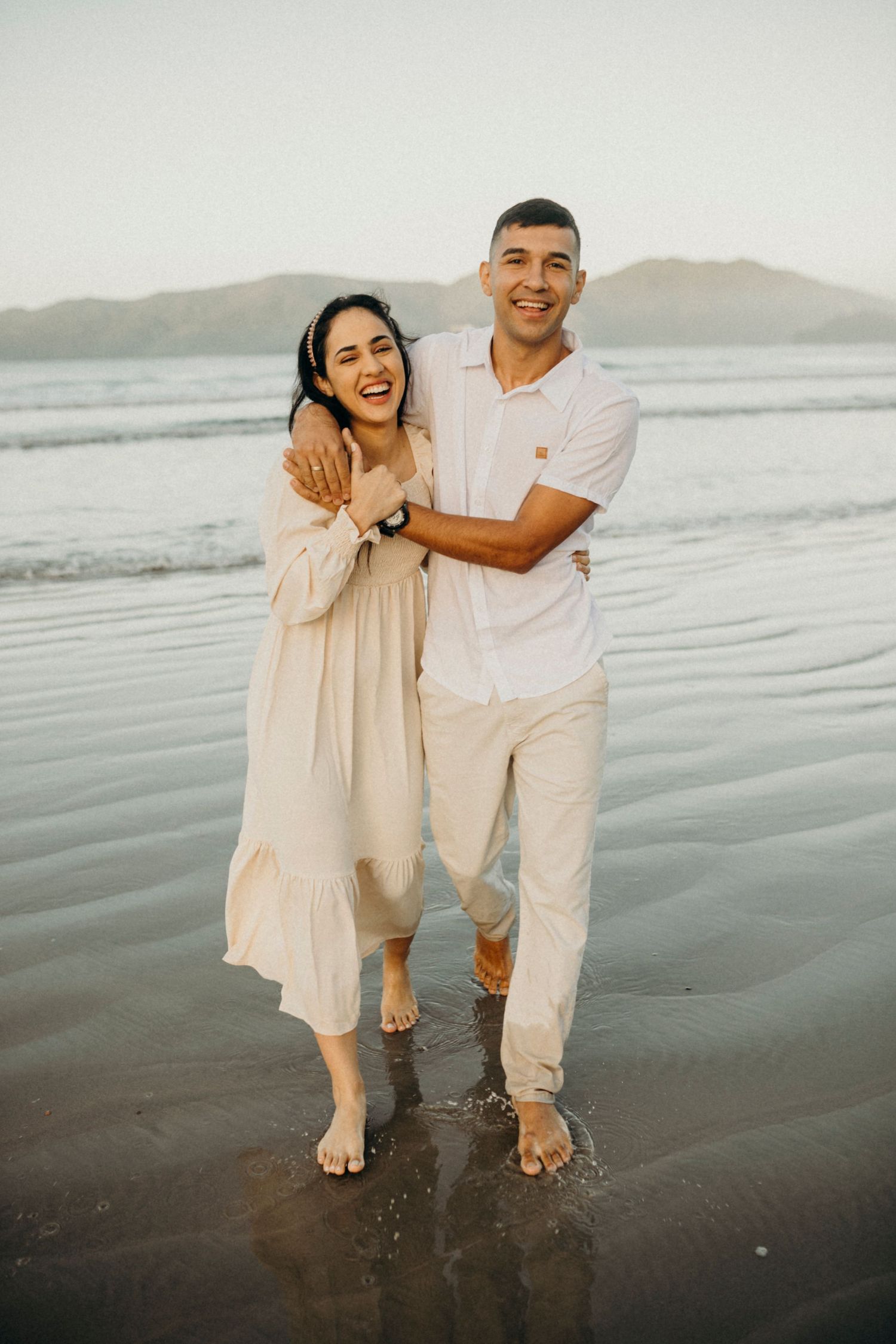 Couple laughing together on a beach at sunset wearing white and cream colored outfits.