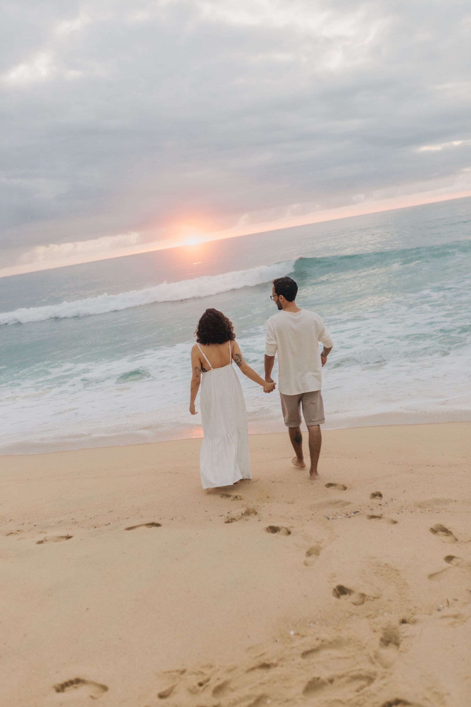 A couple walks hand in hand on beach at sunset.