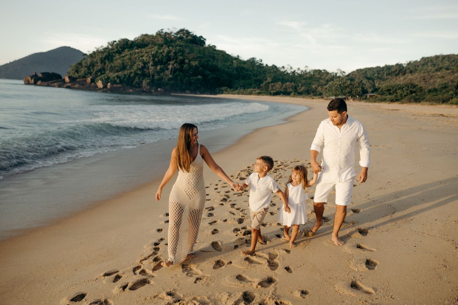 Family walking along beach shore leaving footprints in sand during sunset.