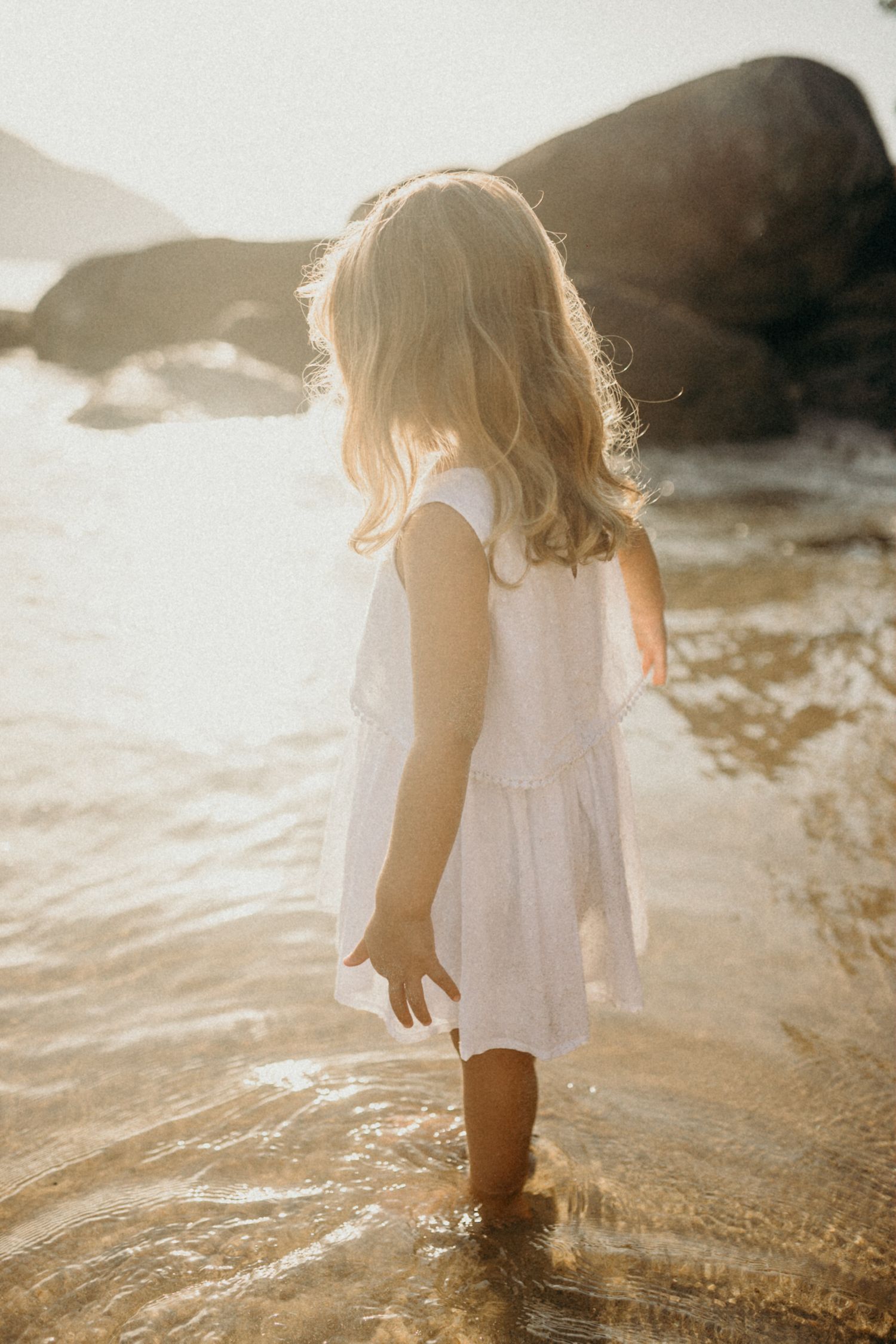 Small child in white dress splashing in gentle ocean waves at sunset with golden light.