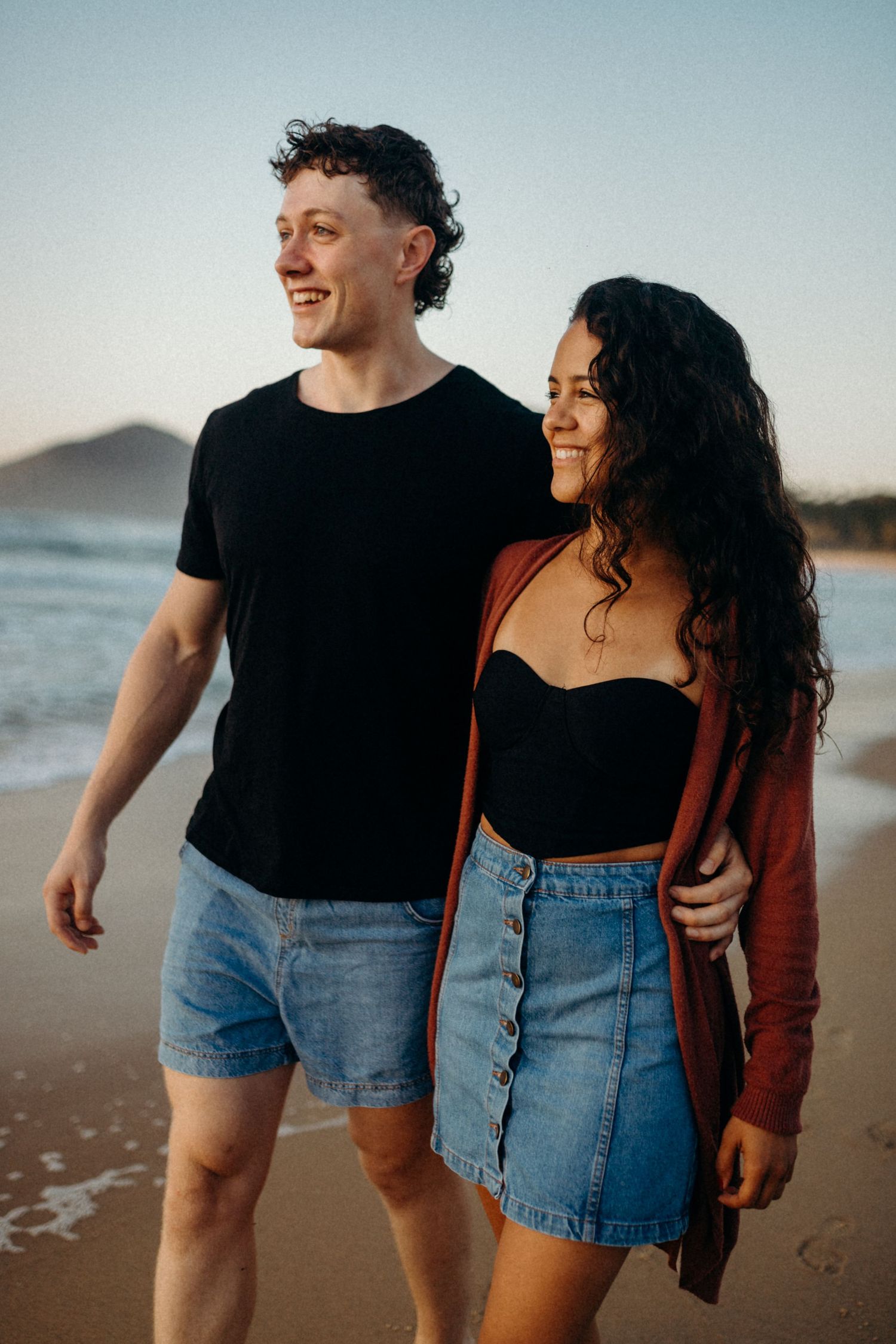 Happy couple walking together on beach during sunset with coastal scenery.