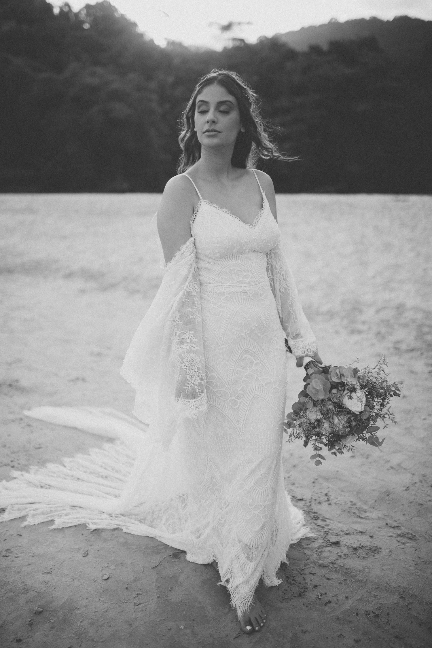 Ethereal black and white bridal portrait shows a flowing lace dress and bouquet against a beach backdrop.