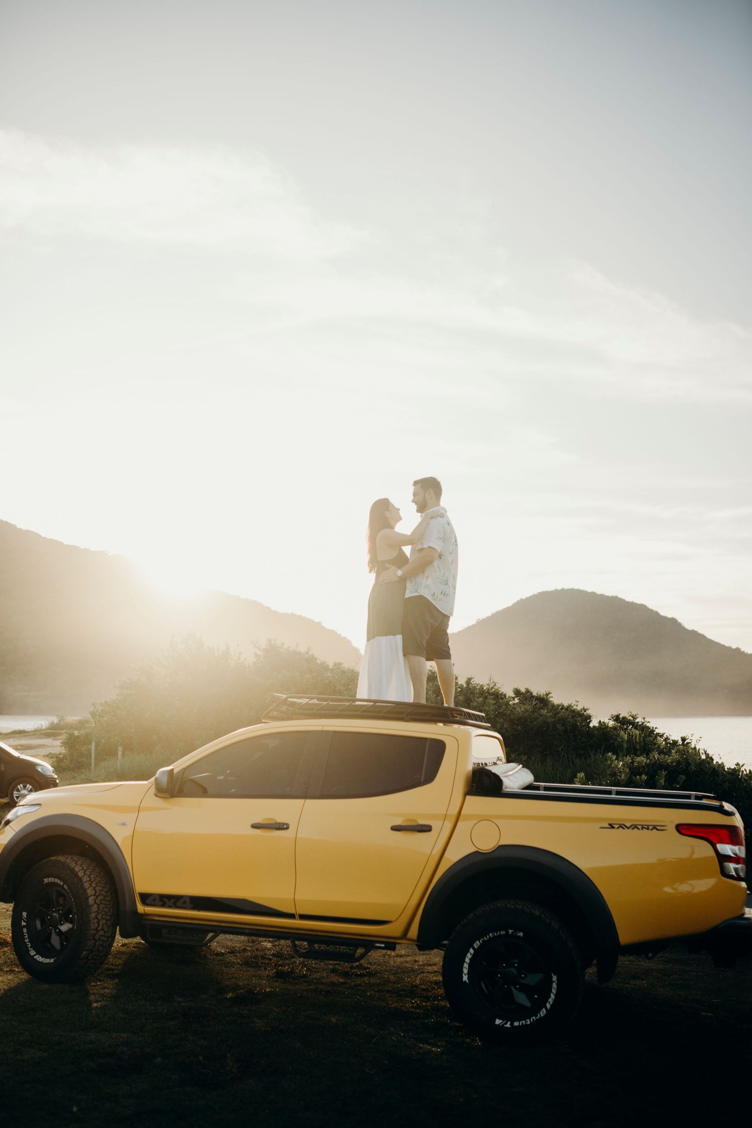 Couple embracing near a yellow pickup truck at sunset with ocean views.