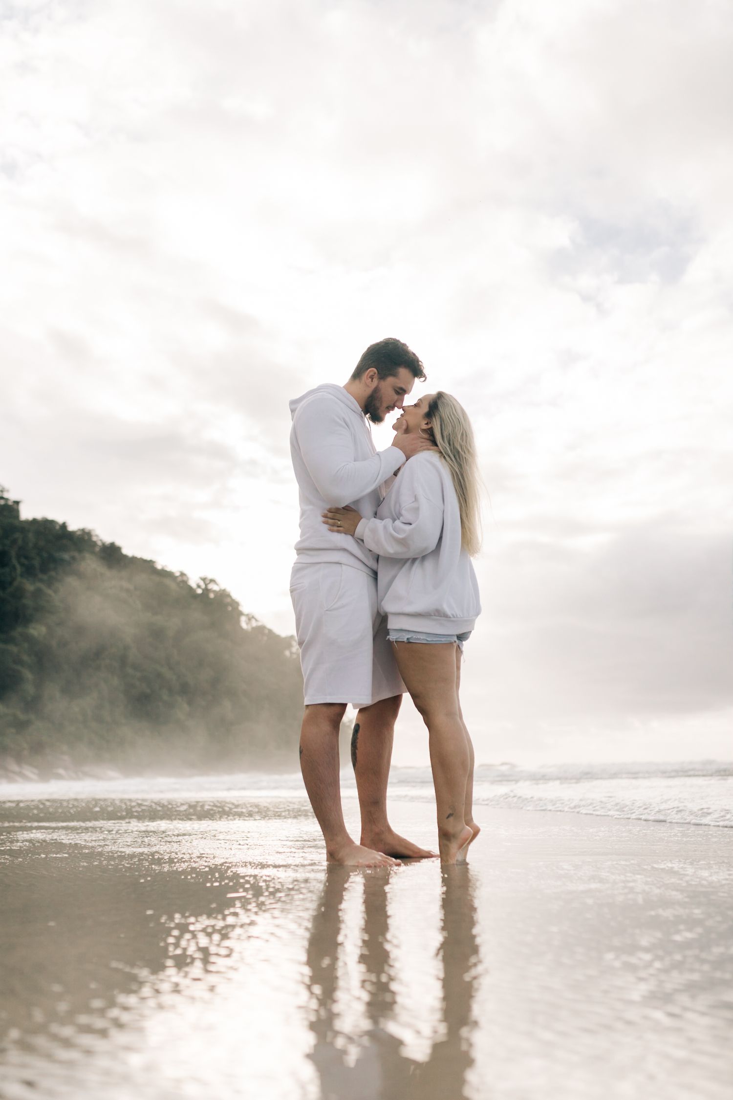 A romantic couple embraces on a foggy beach shoreline wearing coordinated white outfits.