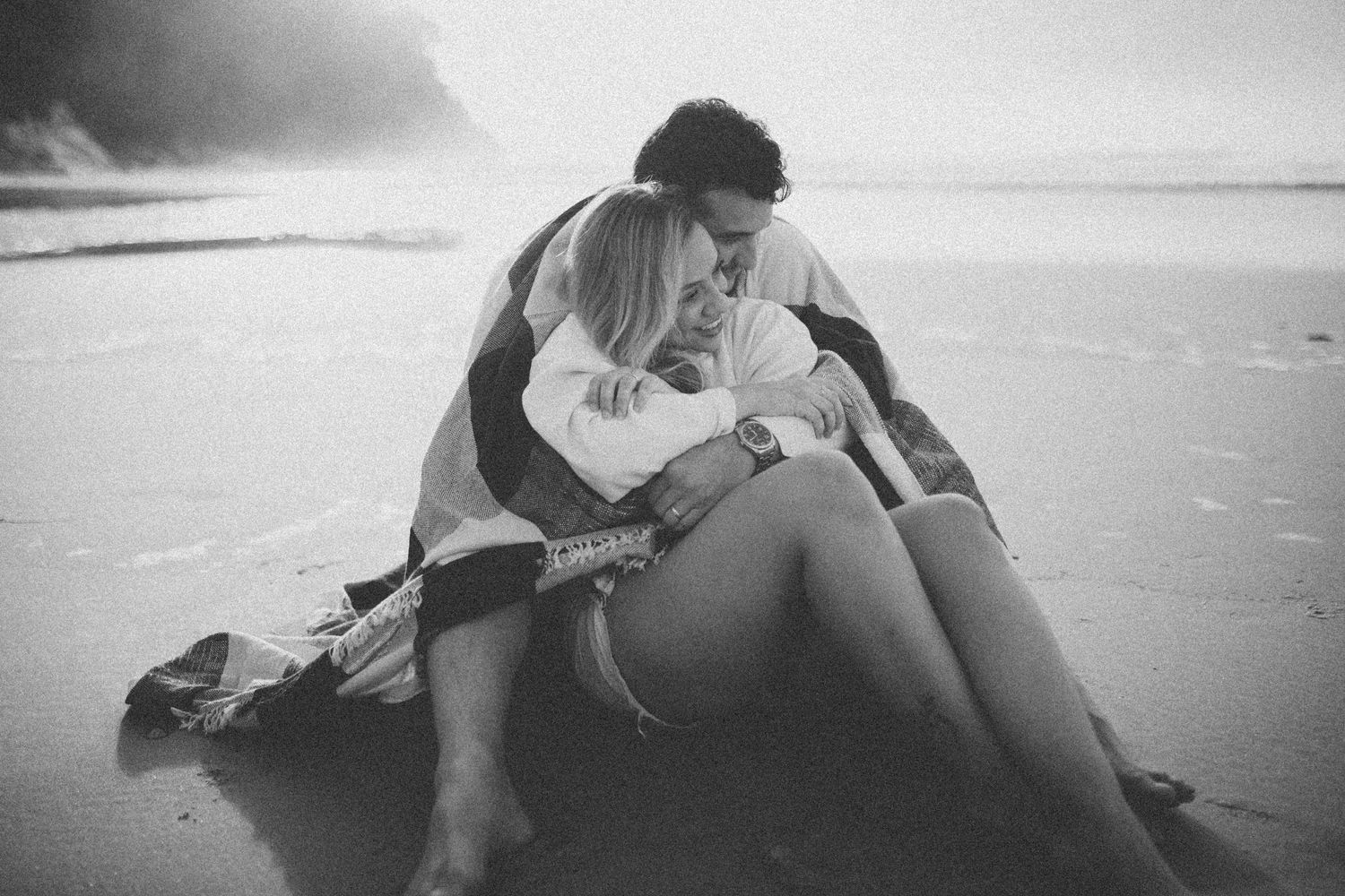 An intimate black and white photo of a couple cuddling on the sandy beach.