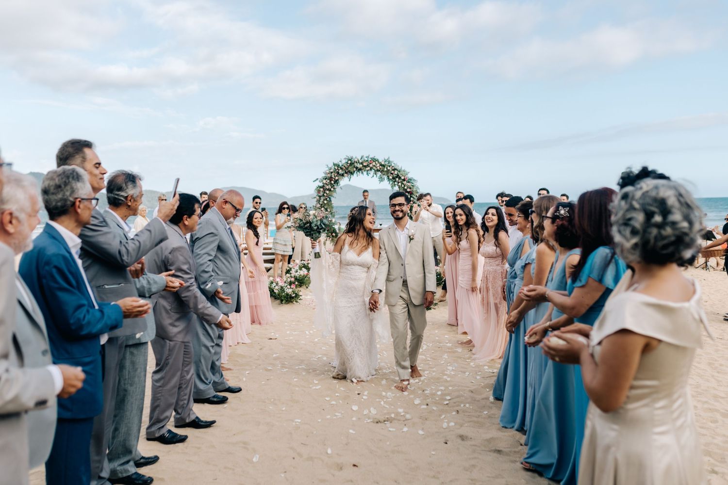 Wedding guests in blue and grey attire form an aisle on the beach during the ceremony exit.
