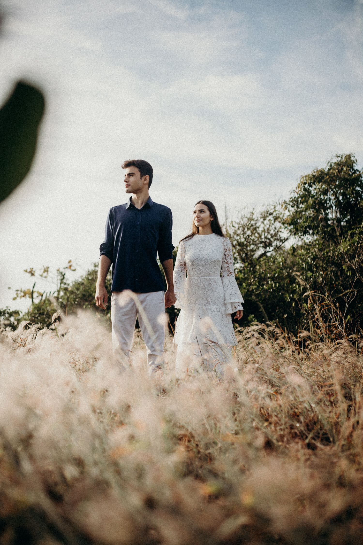 Couple in formal wear walking through golden grass field on sunny day.