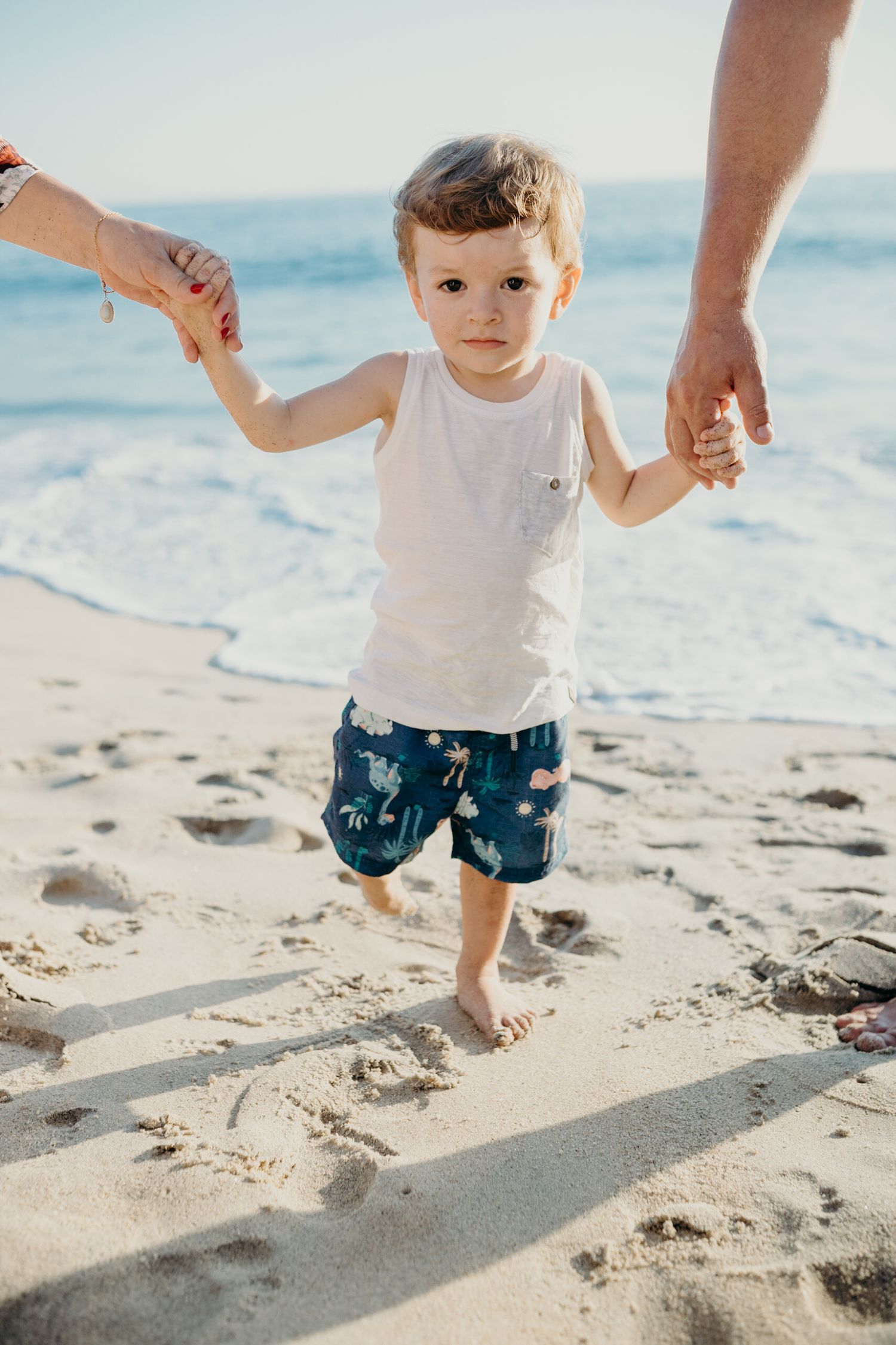 Family holds hands with toddler walking along sunny beach shoreline.