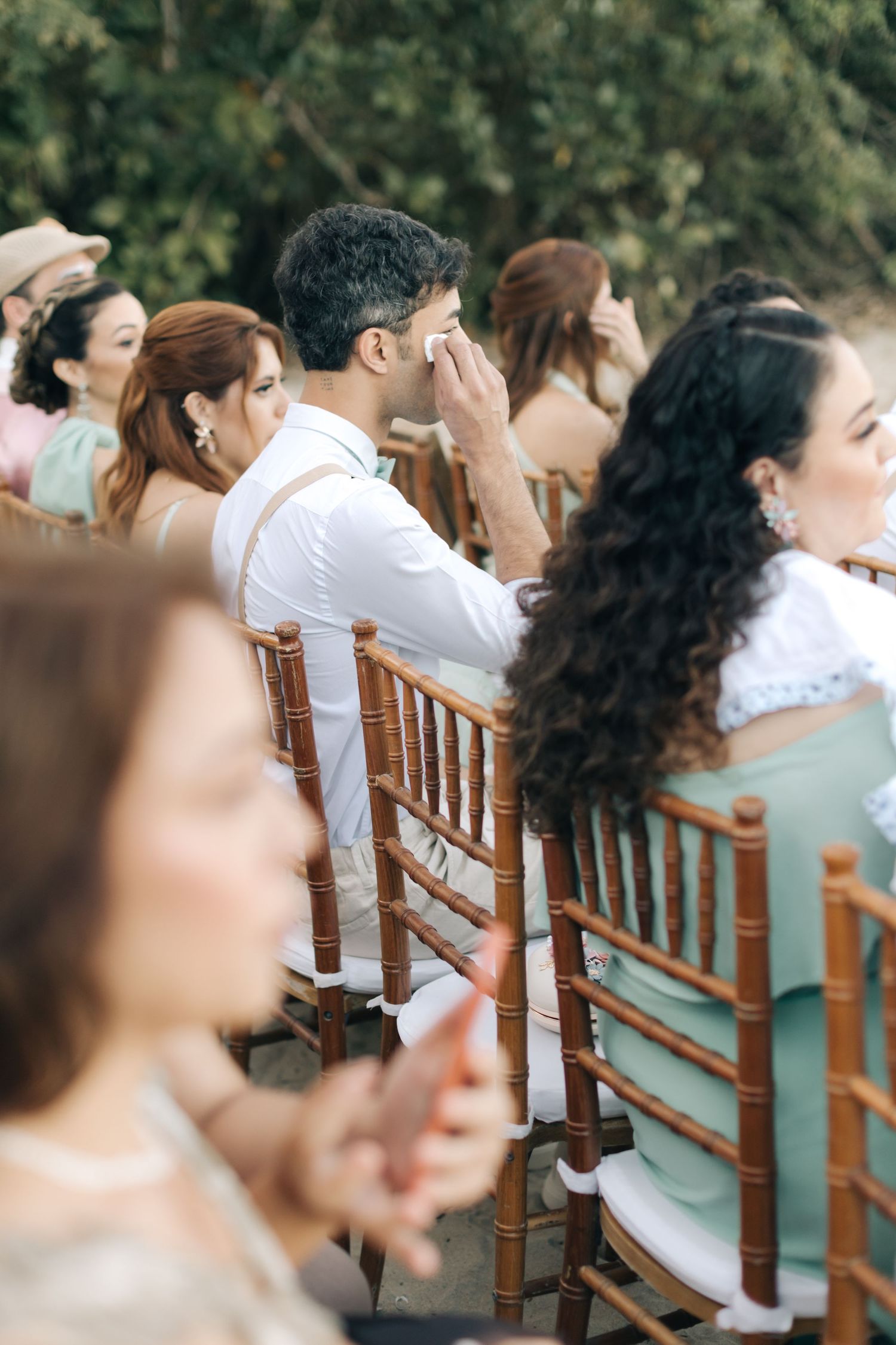 Wedding guests seated in wooden chiavari chairs react emotionally during an outdoor ceremony.